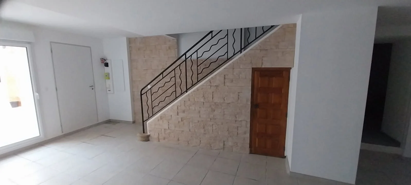 Intérieur d'une pièce avec un escalier en métal et mur en briques, porte en bois, et fenêtres laissant entrer de la lumière naturelle.