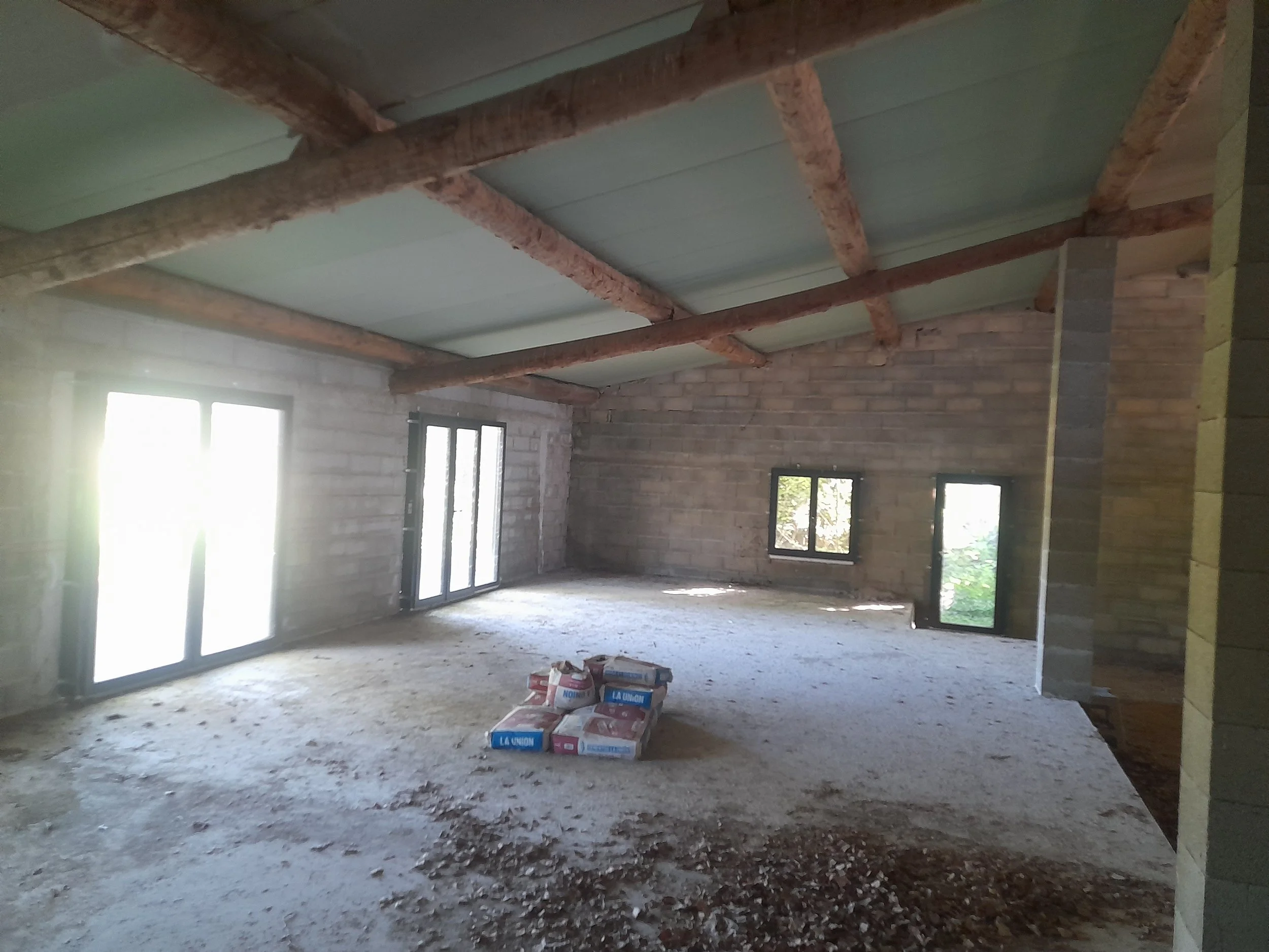 Une pièce en construction avec un plafond en cours d'installation, des murs en briques et plusieurs fenêtres laissant entrer de la lumière naturelle, avec des sacs de ciment et des débris au sol.