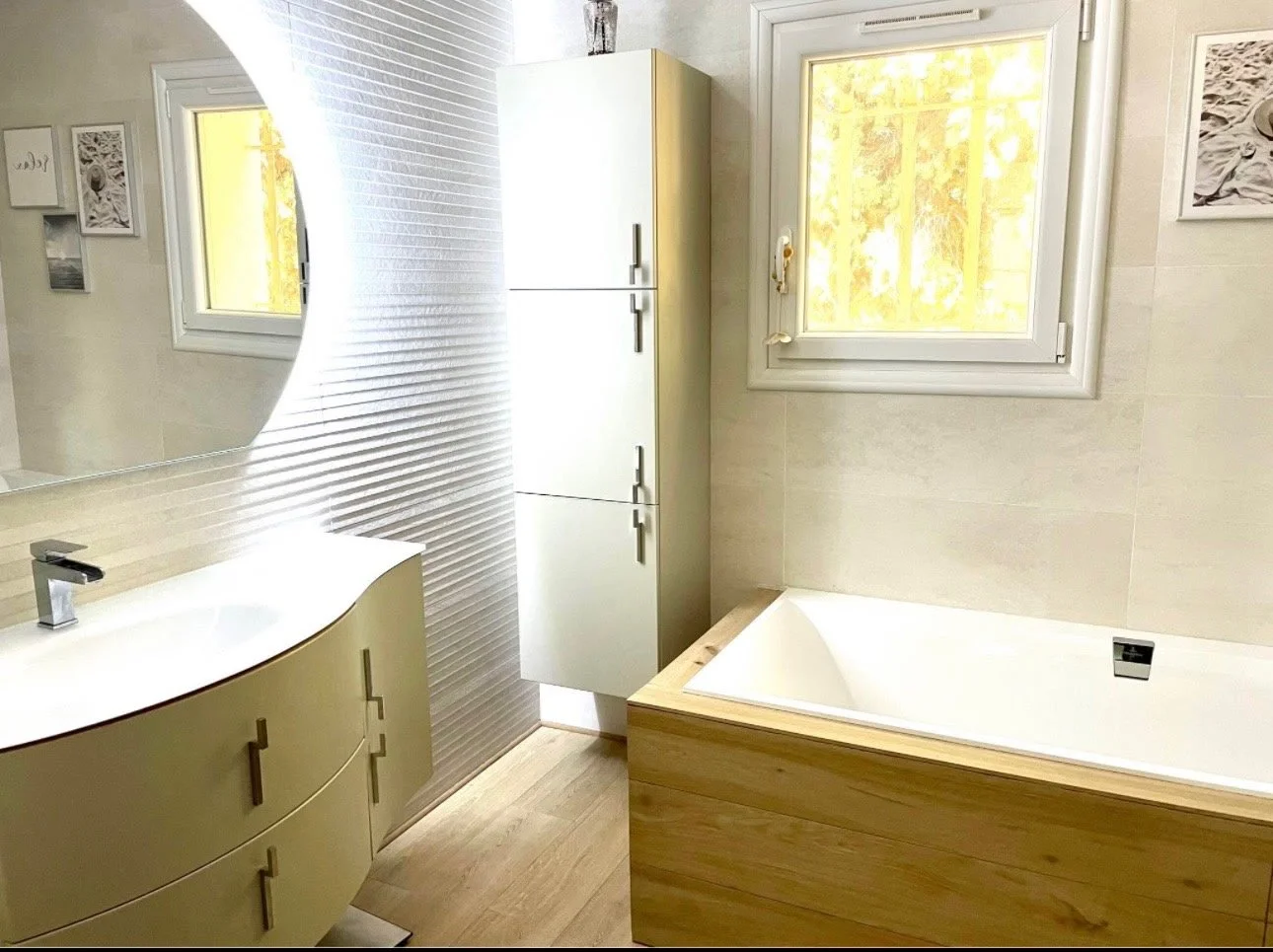 Salle de bain moderne avec une baignoire encastrée en bois, un lavabo avec un meuble de rangement beige, une grande fenêtre laissant entrer la lumière naturelle, et quelques tableaux décoratifs sur le mur.