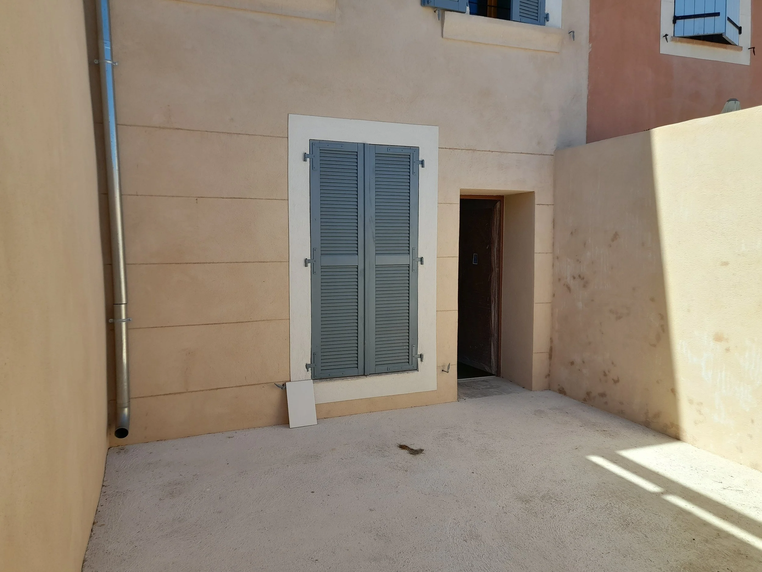 Mur beige avec deux fenêtres à volets bleus et une porte entrouverte. Canalisations en métal le long du mur et sol en béton. Ombres portées sur le sol.