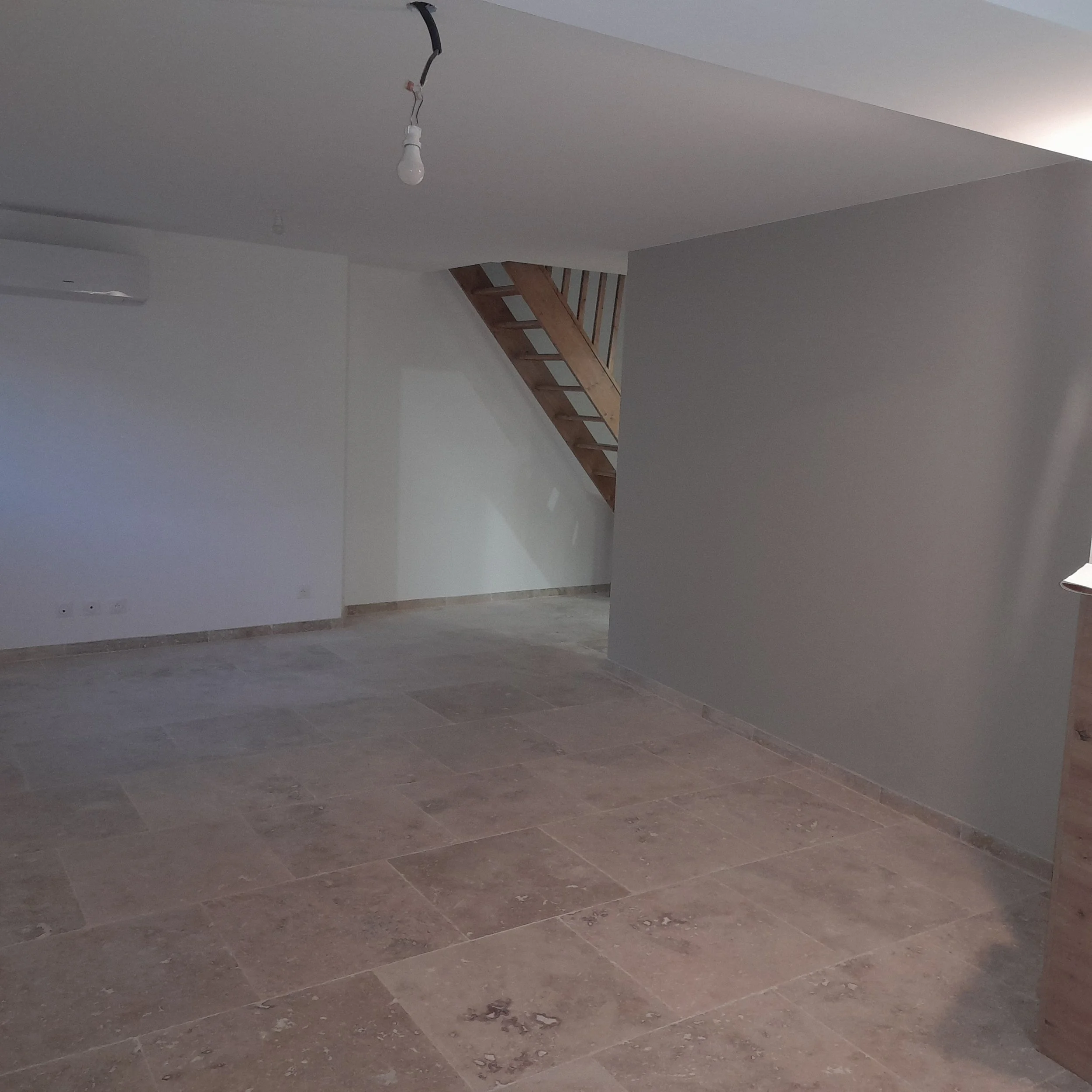 Salle en construction avec des murs blancs, un plafond sans peinture et un escalier en bois menant à un étage supérieur, lumière naturelle