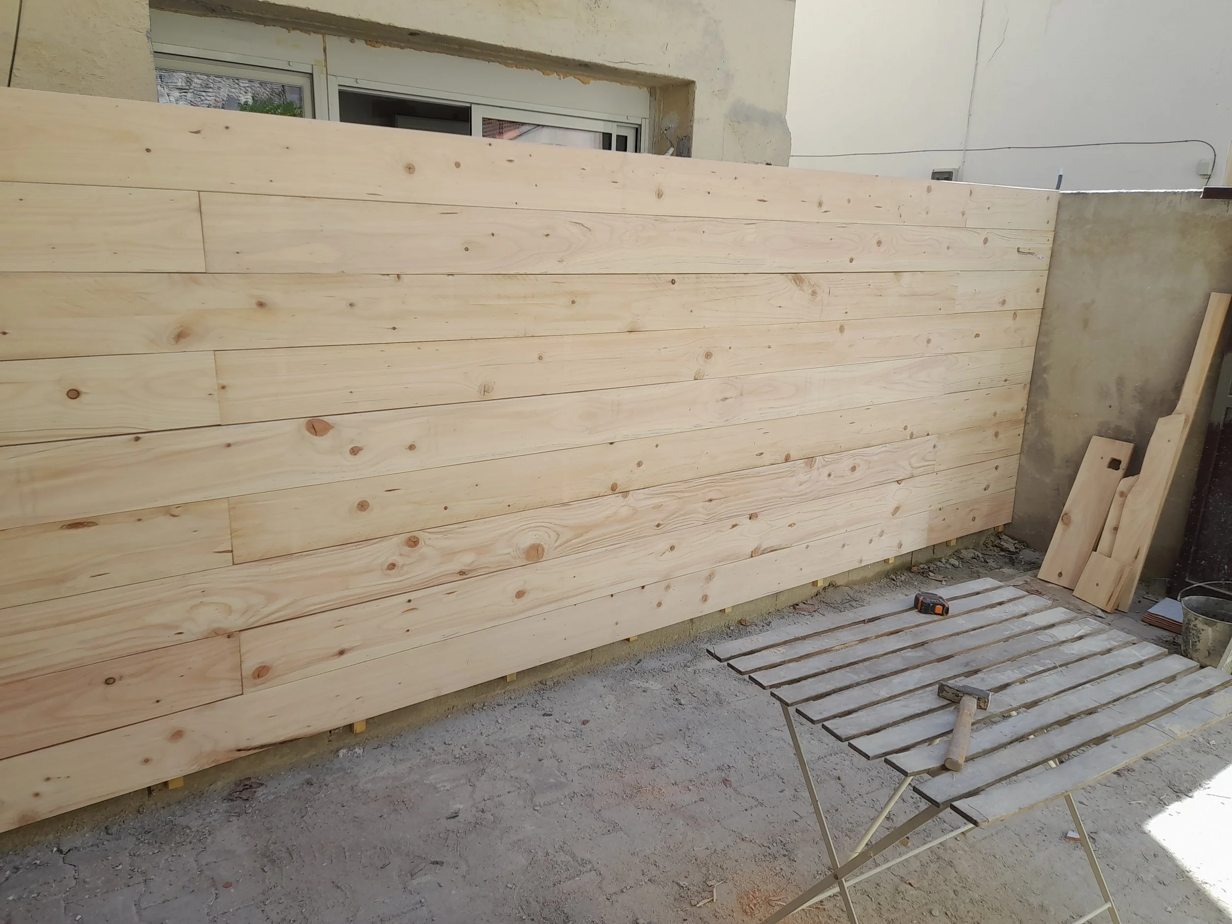 Un mur en bois en cours de construction dans un espace intérieur, avec des planches de bois claires empilées horizontalement.
