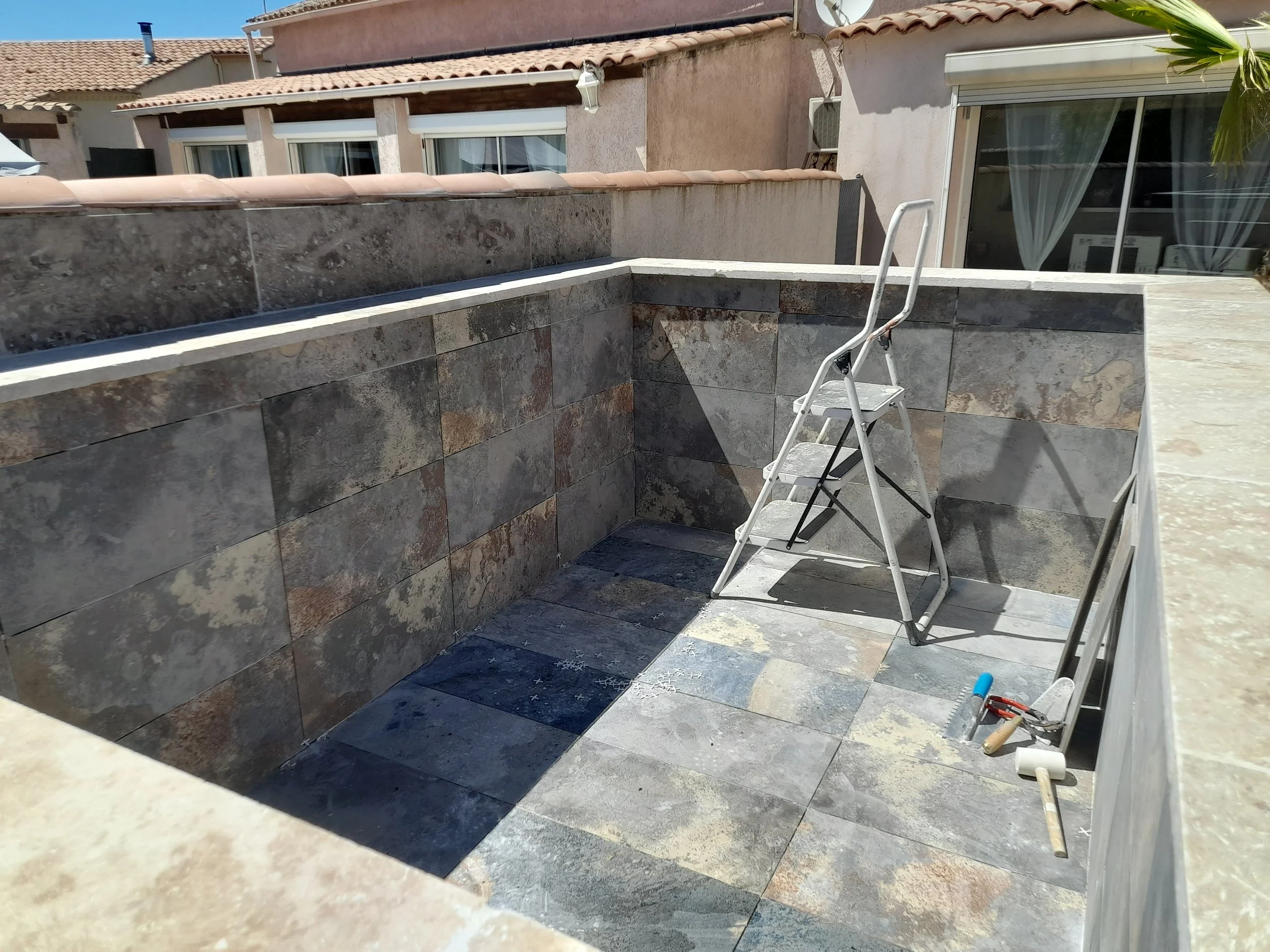 Balcon en cours de rénovation avec des carreaux de pierre, une échelle pliante blanche et des outils, surplombant des maisons résidentielle avec des fenêtres avec rideaux blancs.