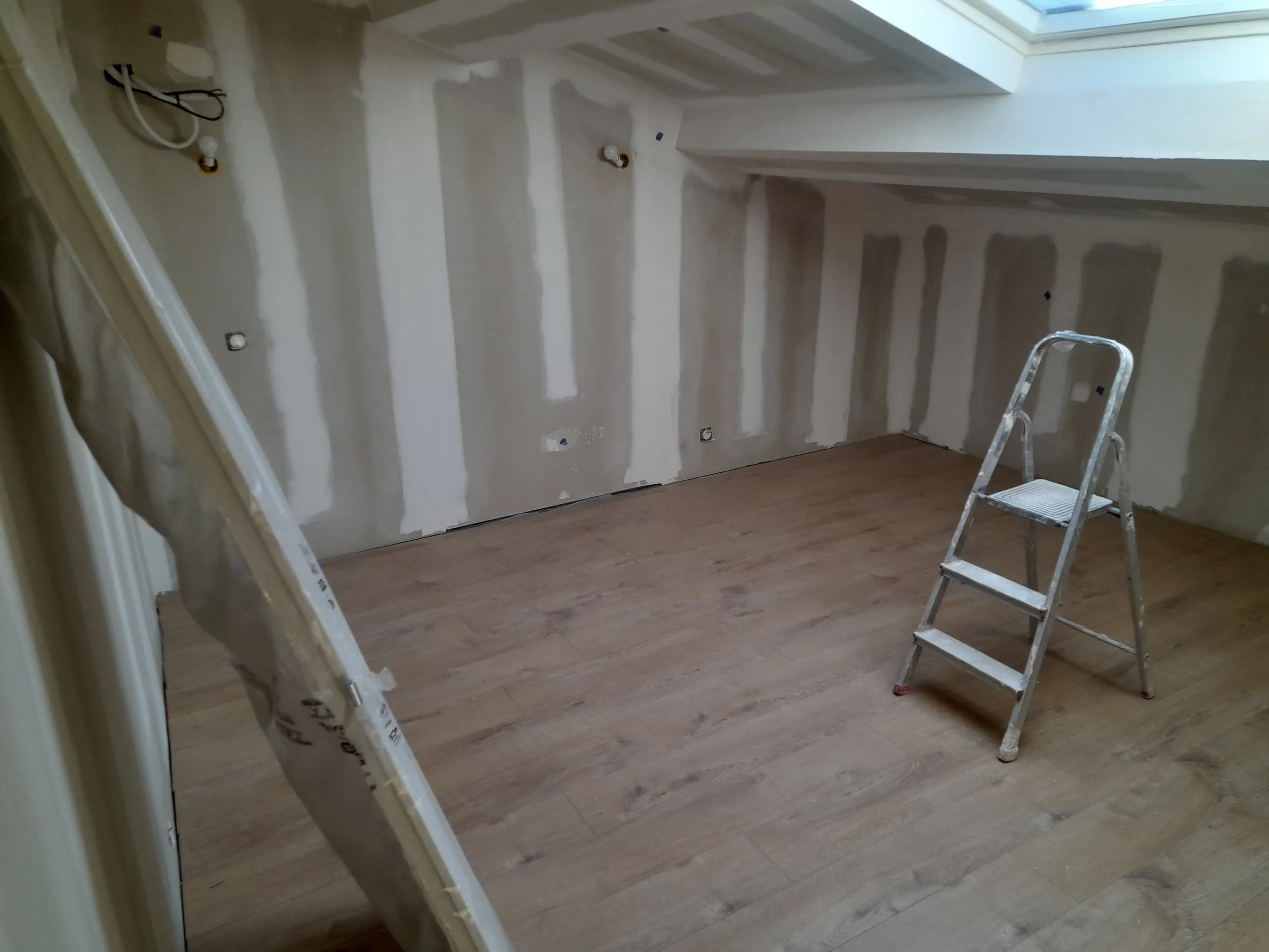 Une pièce en cours de rénovation avec murs en plâtre en train d'être préparés. Il y a deux escabeaux en métal, un à gauche et un autre à droite, ainsi qu'un sol en bois. Une fenêtre au plafond laisse entrer la lumière naturelle.