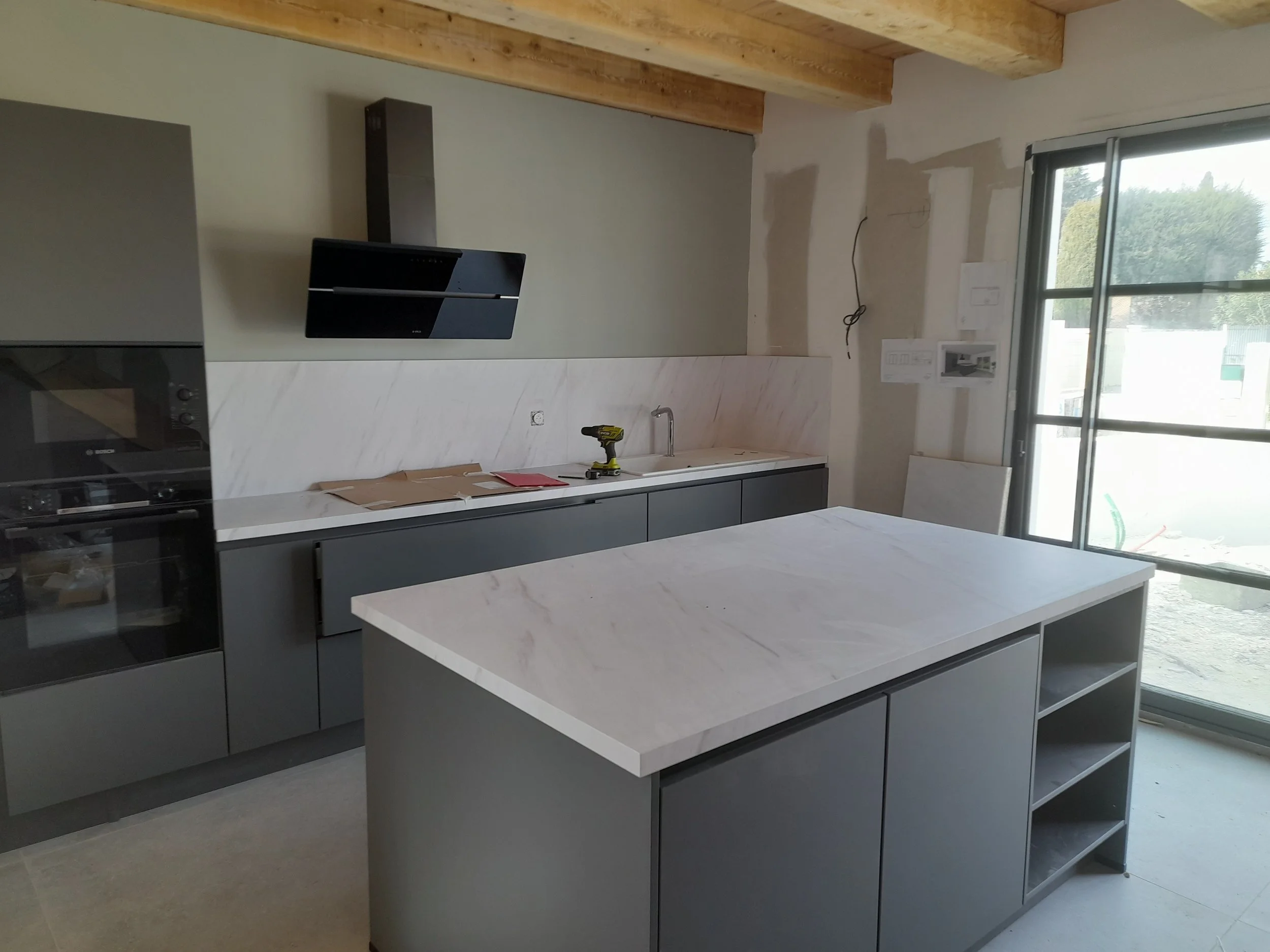 Une cuisine en cours de rénovation, avec des plans et un tournevis sur le comptoir. La pièce a de grands fenêtres laissant entrer la lumière naturelle.