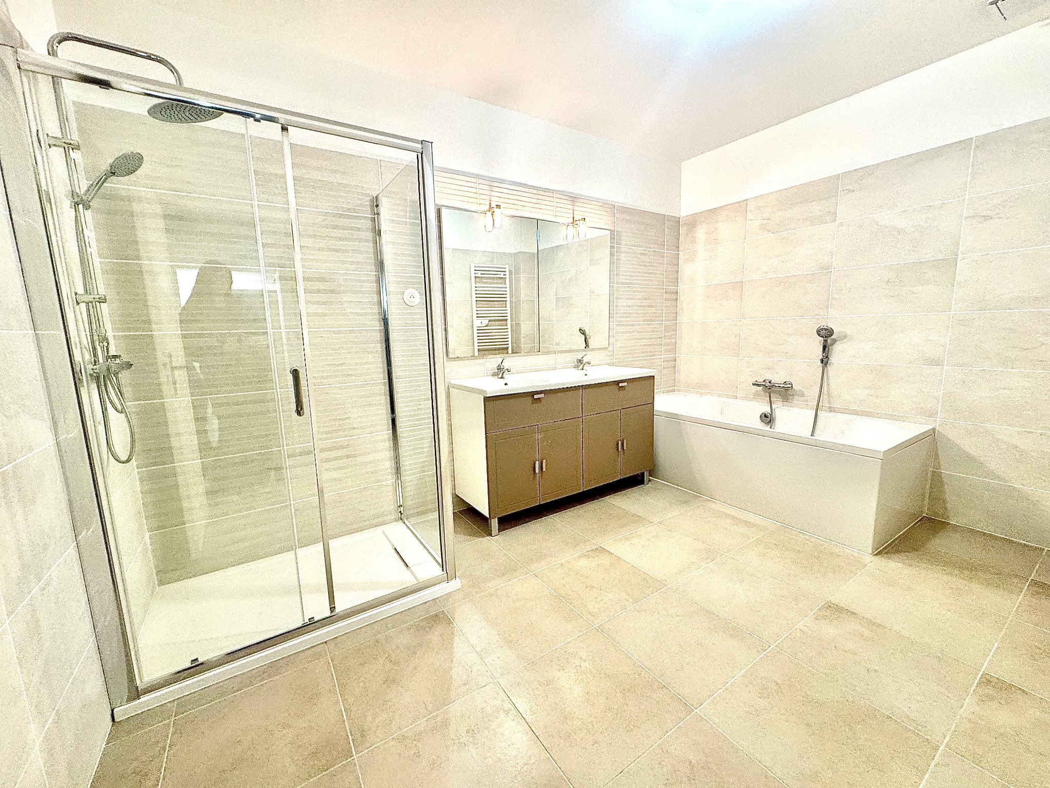 Une salle de bain moderne avec une cabine de douche, une baignoire, un miroir et un meuble double lavabo. Les murs et le sol sont carrelés de beige. La salle est bien éclairée.