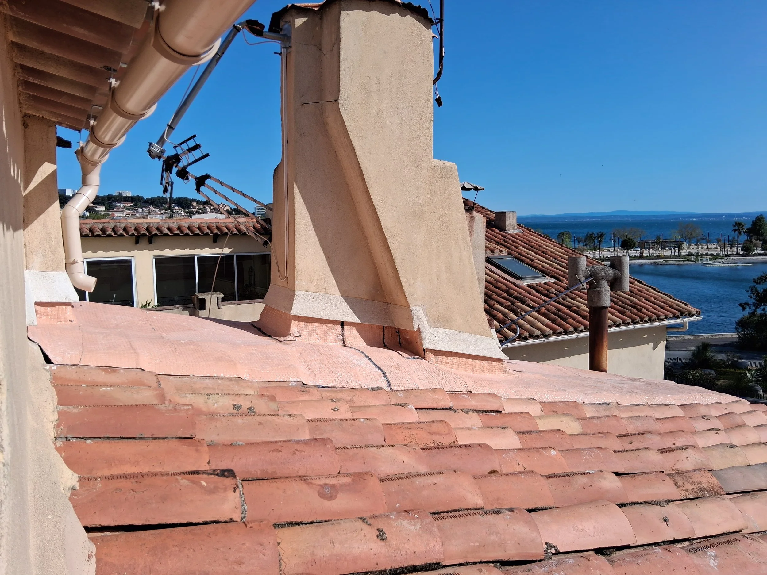 Vue d'un toit avec des tuiles en terre cuite, des tuyaux de plomberie et une cheminée, avec en arrière-plan la mer et un ciel bleu clair.