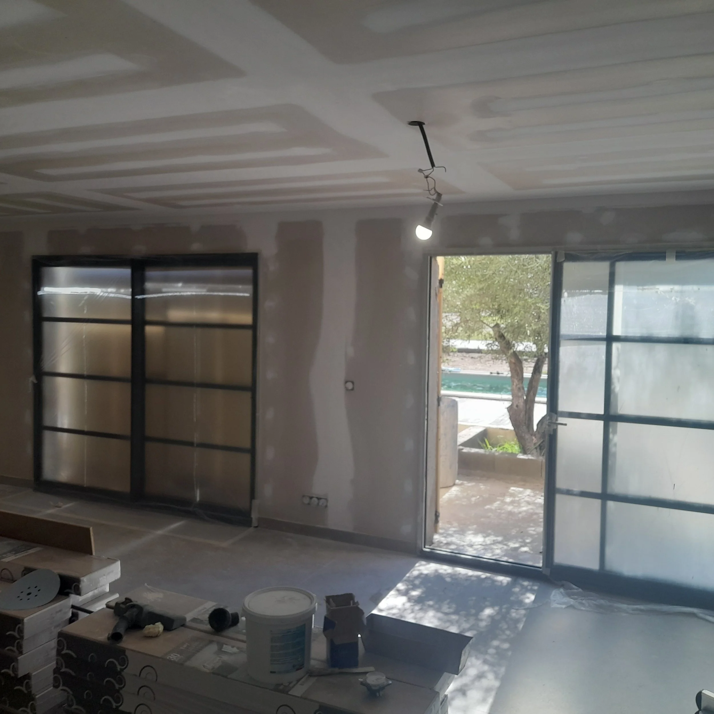 Intérieur d'une pièce en construction avec plafond suspendu en cours d'installation, éclairage suspendu, outils et matériaux de construction sur le sol et une porte donnant sur l'extérieur.