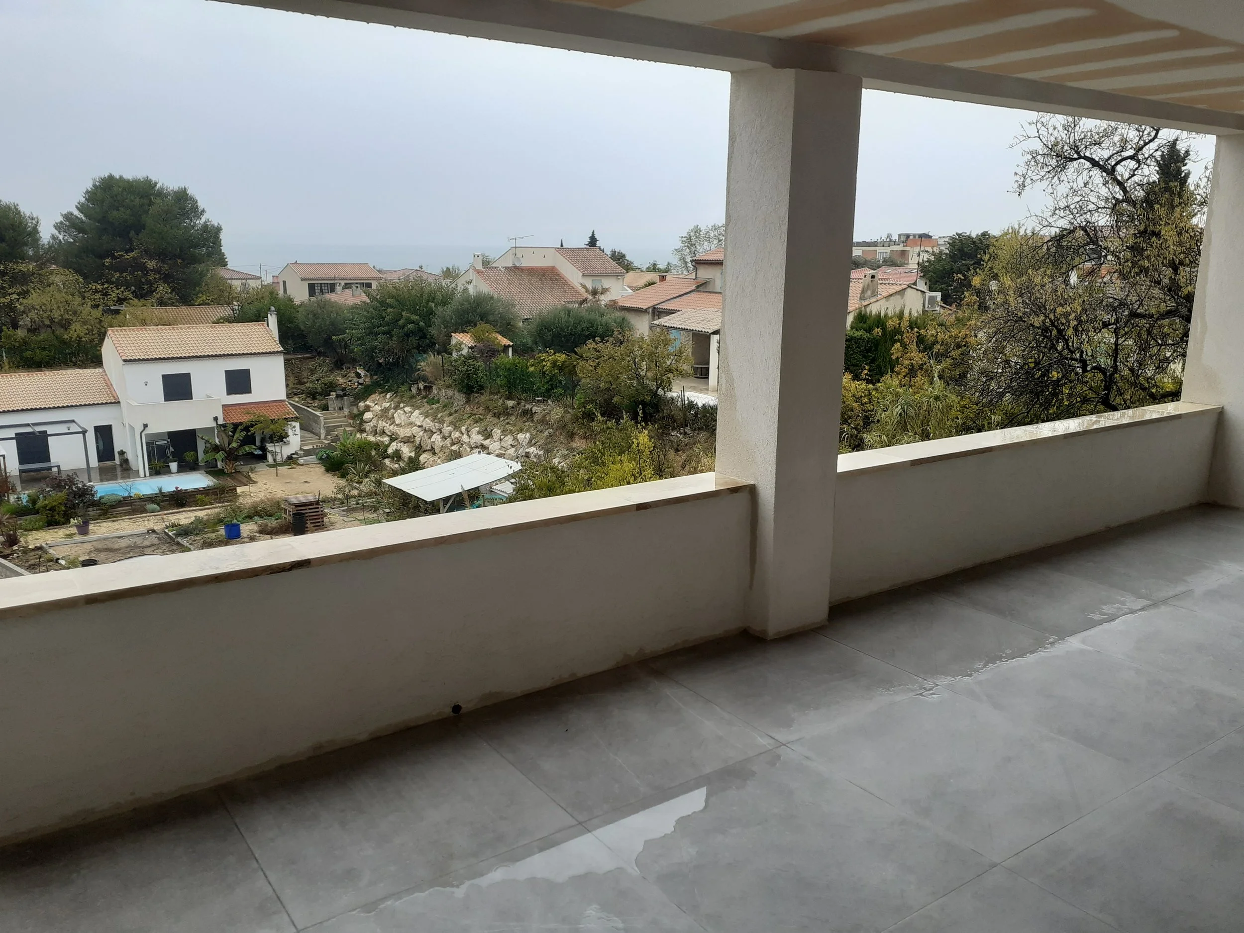 Vue d'une terrasse partiellement couverte surplombant des maisons avec des toits en tuiles dans un quartier résidentiel calme. La scène semble être un jour nuageux ou brumeux.