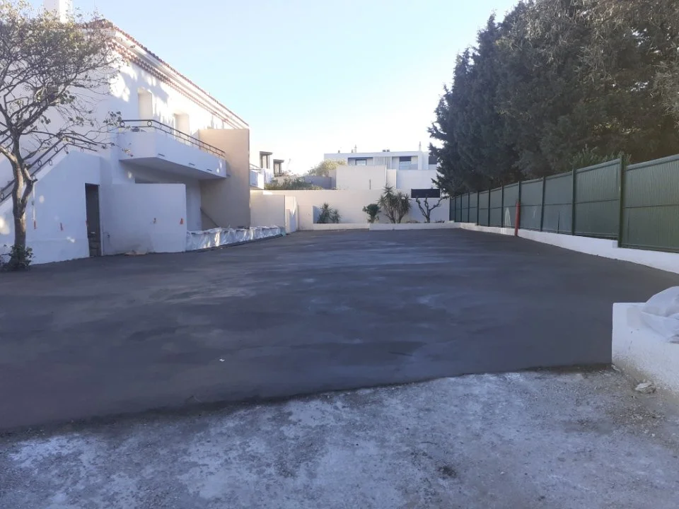 Une zone de stationnement extérieure récemment asphaltée, entourée de murs blancs et d'une clôture verte, avec des arbres et un bâtiment en arrière-plan.