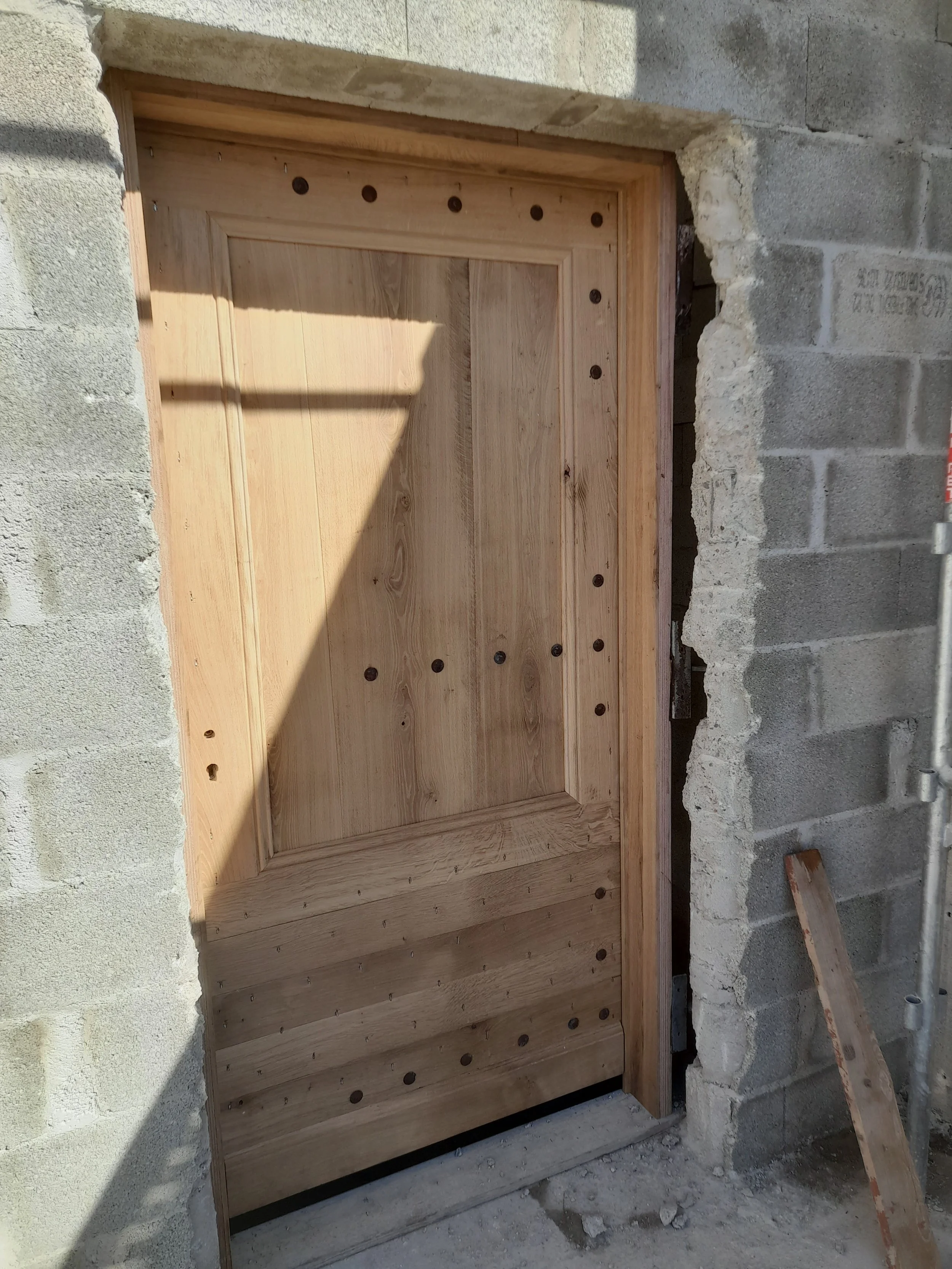 Porte en bois en cours d'installation dans un mur en pierre. La porte est en bois clair avec des trous pour la fixation. Le mur autour est en pierre brut, en construction.