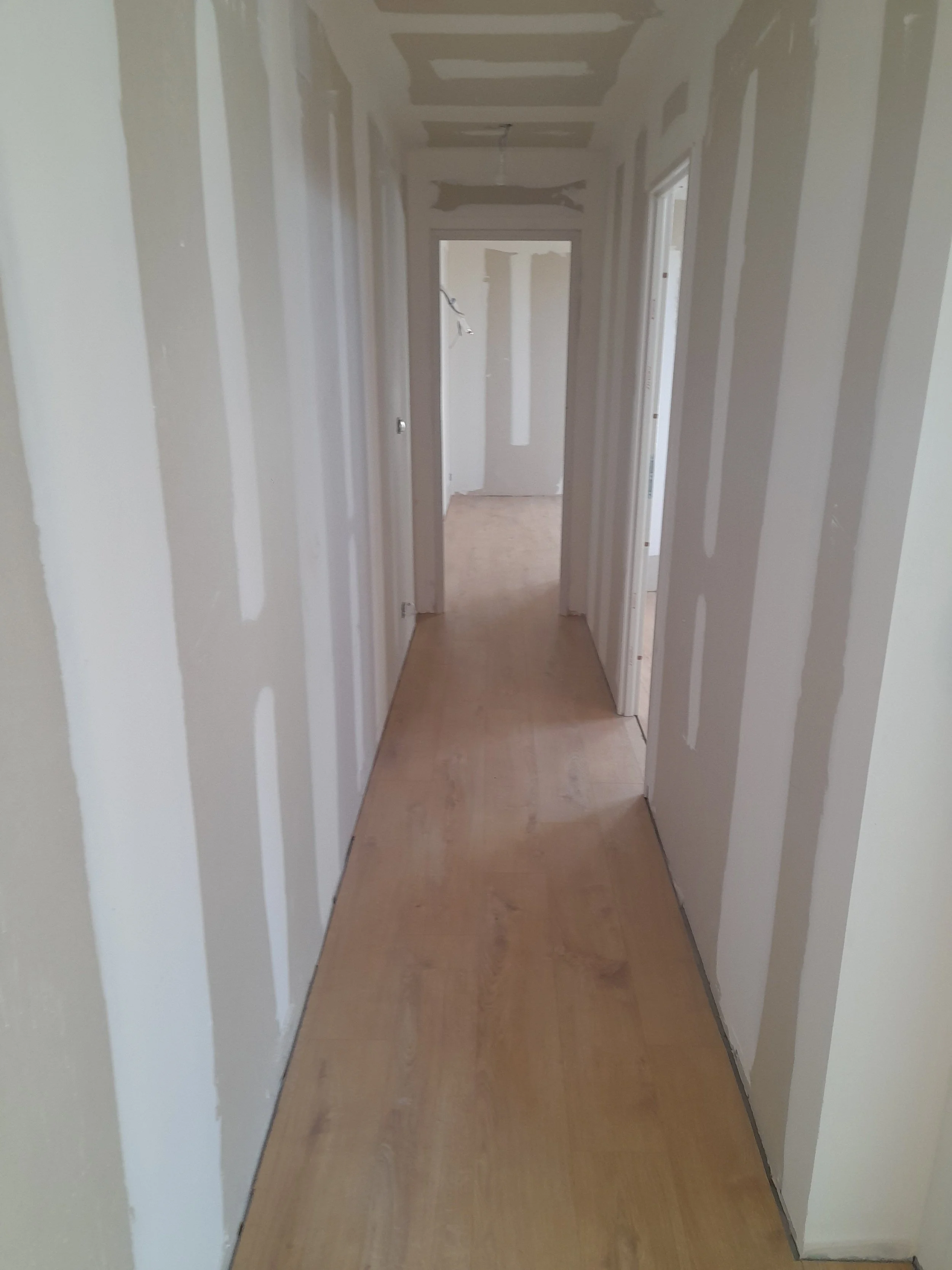 Un couloir en cours de rénovation avec des murs en plaques de plâtre, encore à peindre ou tapisser, et un sol en bois.