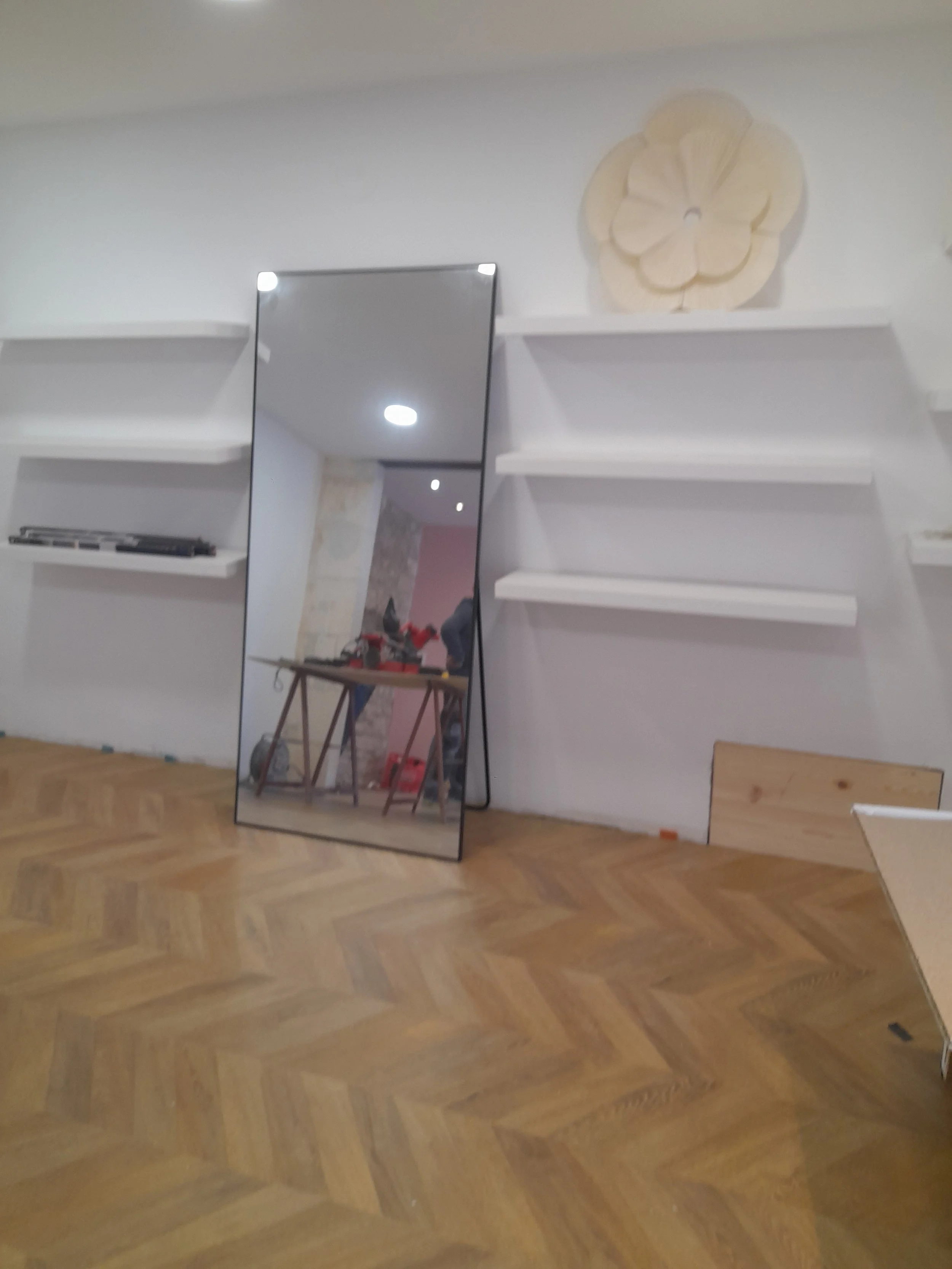 Un grand miroir en verre posé contre un mur blanc, reflétant une table avec des outils et un mur en pierres derrière, avec des étagères blanches vides au-dessus.