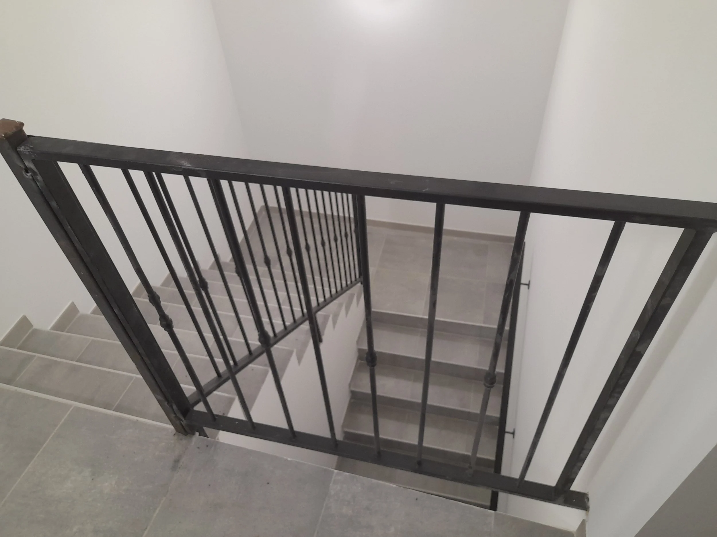 Escalier intérieur avec rampe en métal noir, côtés en blanc, sol en carreaux gris.