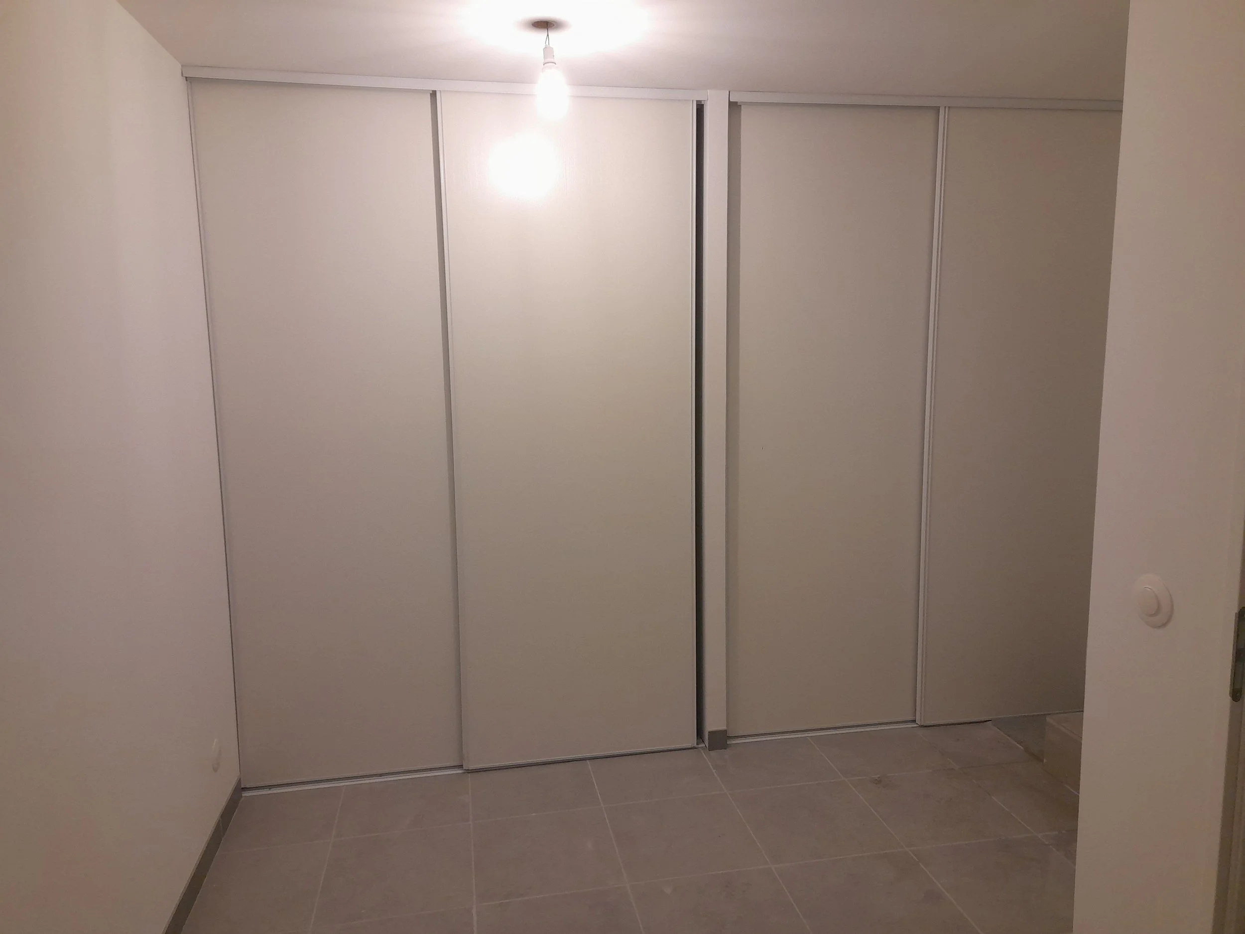 Un grand placard blanc à portes coulissantes dans une pièce avec sol en carrelage gris. Il y a une ampoule allumée suspendue au plafond.