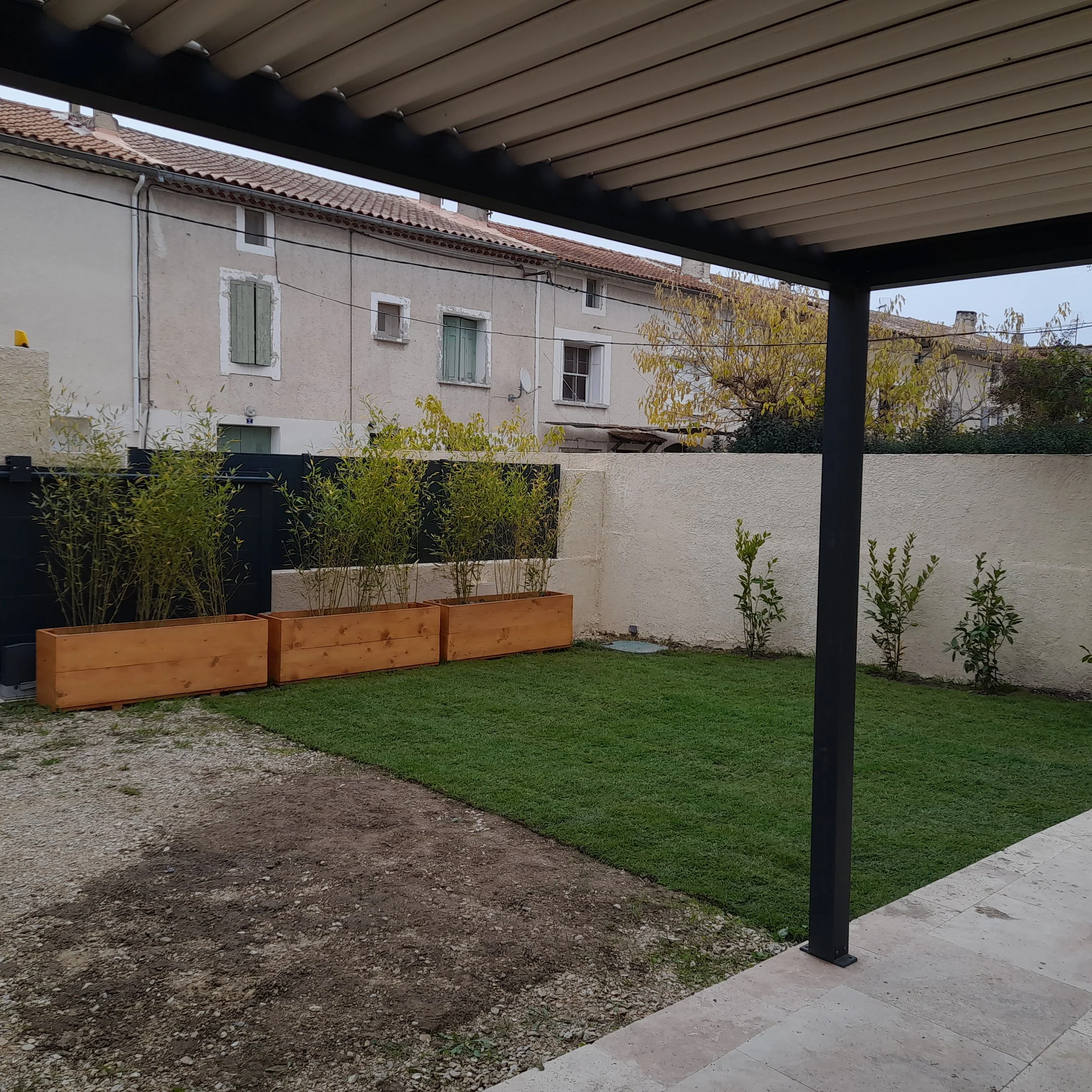 Vue d'un jardin privatif avec un mur en pierre, des plantes en pot en bois, une pelouse, et une pergola avec un plafond en lamelles de bois noir, au-dessus d'une partie de terre et d'une allée en pierre. En arrière-plan, des maisons anciennes avec de