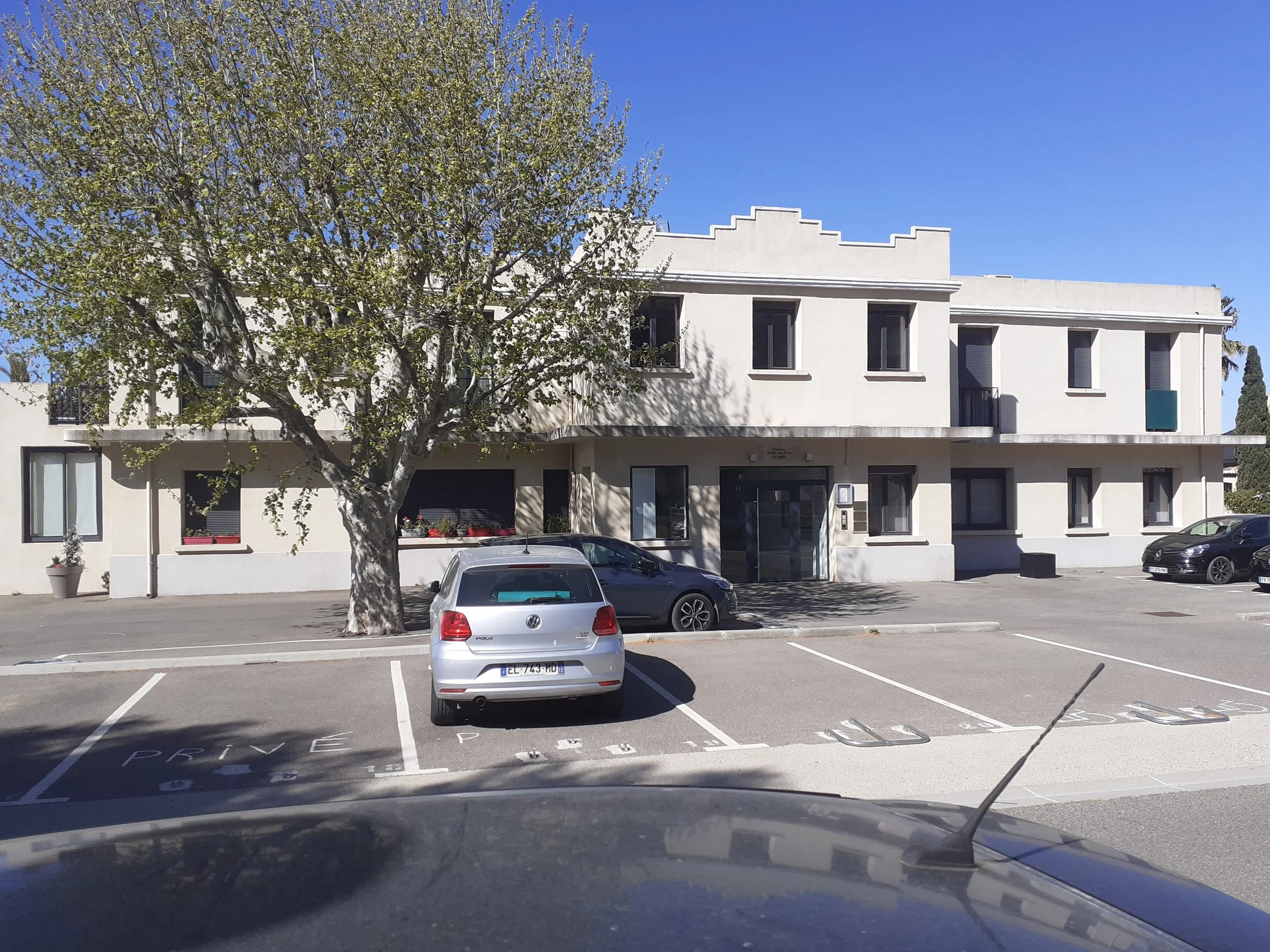Vue depuis l'intérieur d'une voiture d'une place de parking vide devant un immeuble résidentiel blanc avec plusieurs fenêtres, un arbre feuillu et un ciel bleu clair.