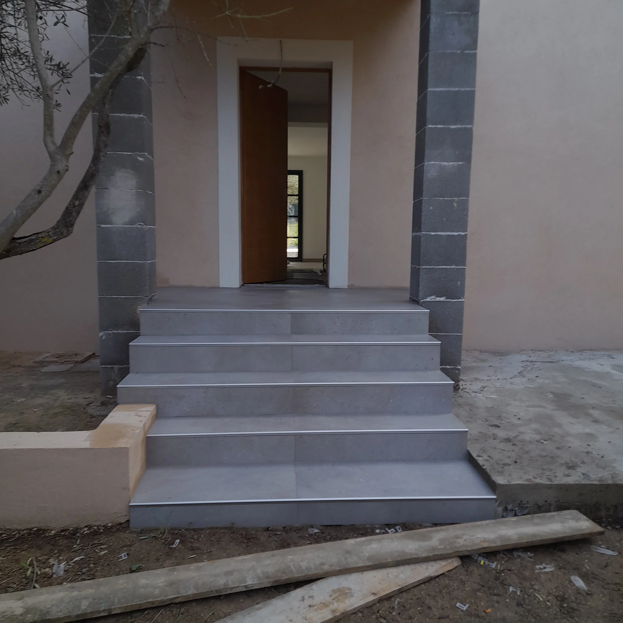 Construction d'une nouvelle maison avec un escalier en pierre, une porte en bois et des murs en béton. Le sol est en terre non pavée.