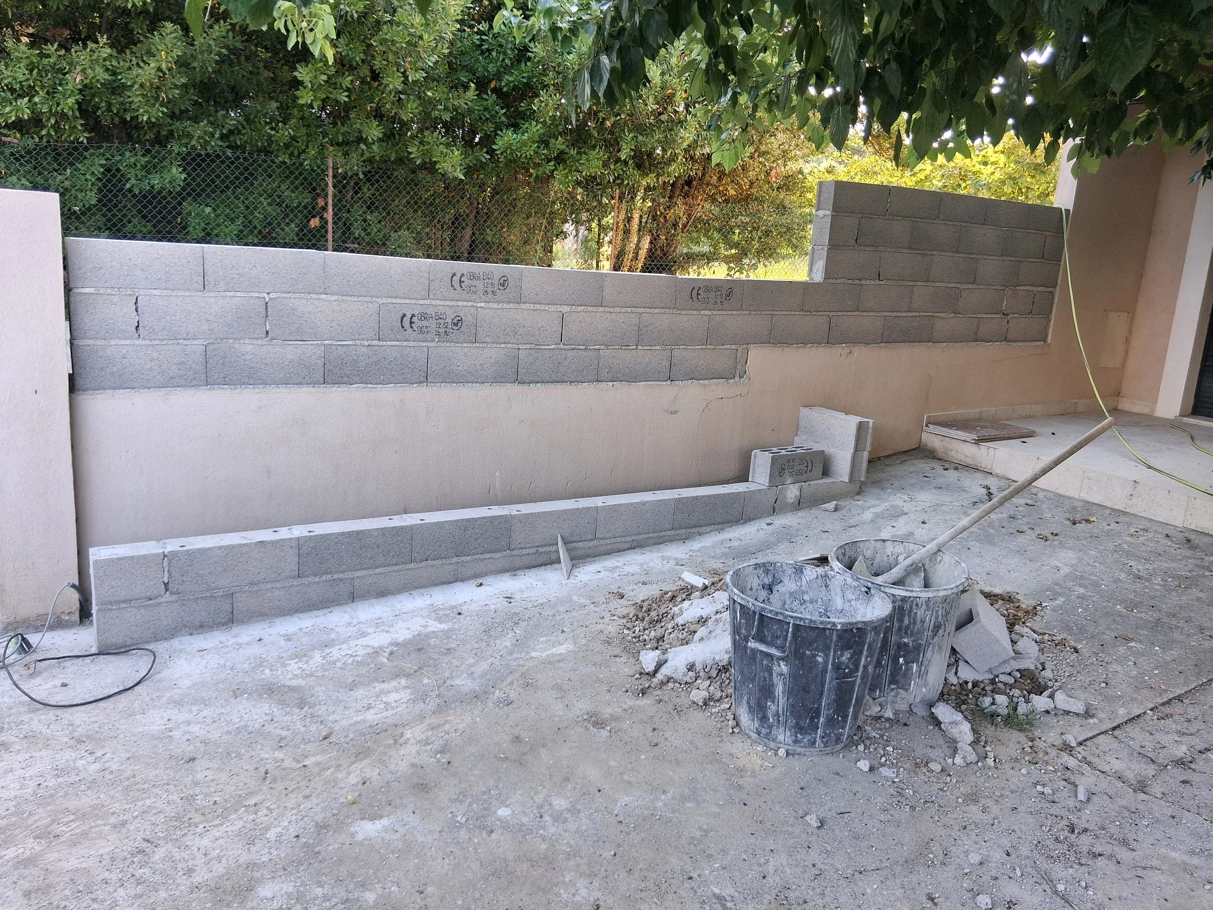 Construction de mur en briques avec bac à charbon et escabeau sur sol en béton, entouré d'arbres et d'une clôture en maillage.
