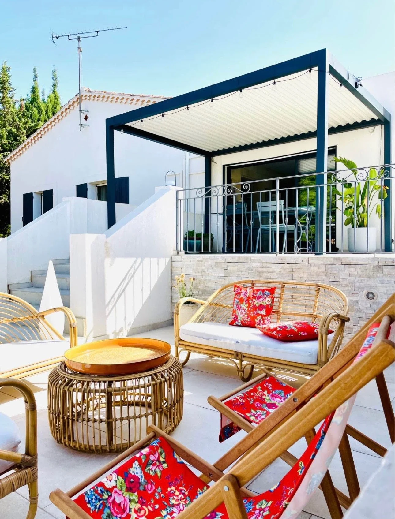 Terrasse extérieure avec mobilier en rotin et coussins colorés, sous un auvent au-dessus d'un escalier menant à une maison blanche moderne avec balcon. Decor végétal avec plantes en pot, ciel bleu en arrière-plan.