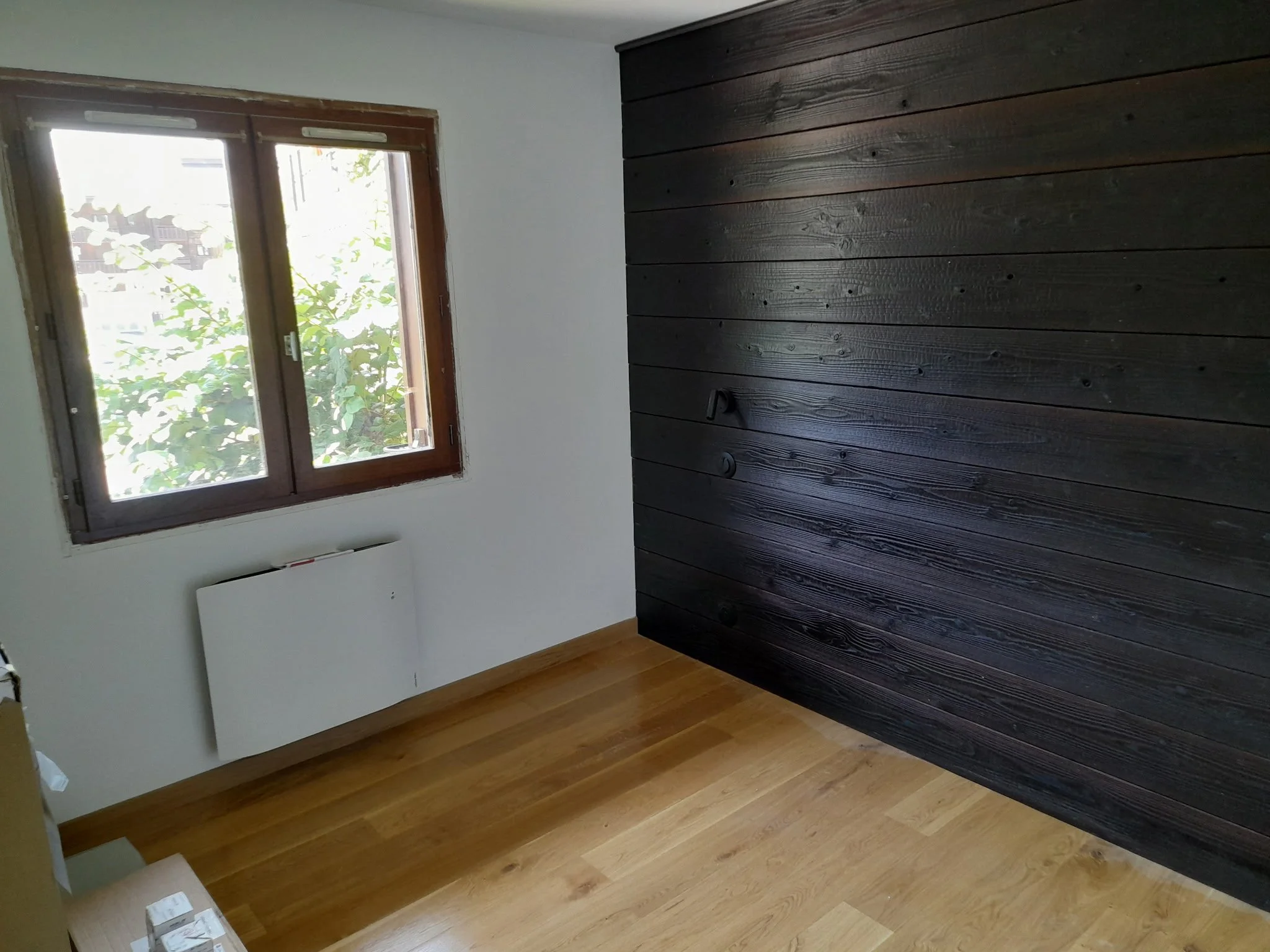 Une pièce avec un mur en bois foncé, une fenêtre laissant passer la lumière du jour, et un chauffage mural blanc.