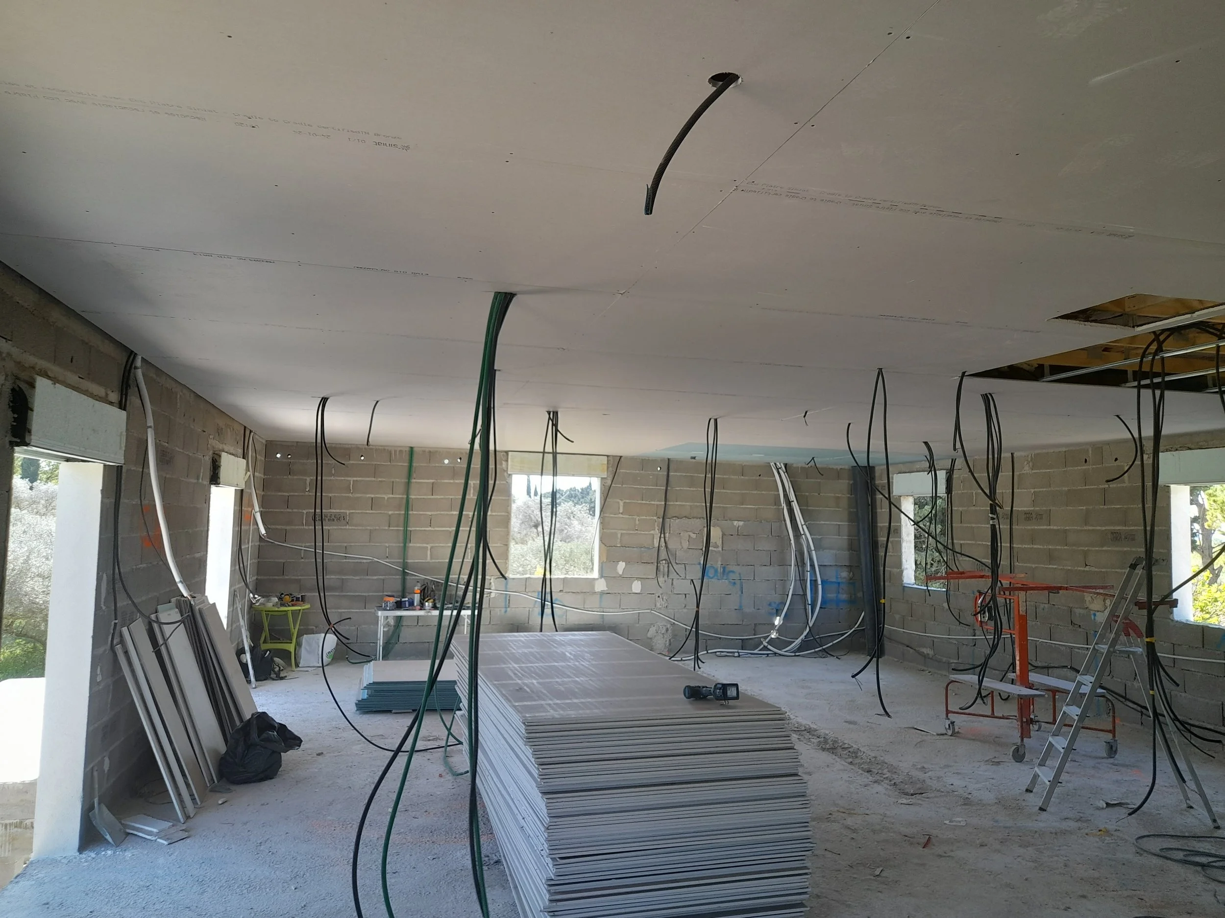 Intérieur d'une construction en cours avec plafond en plaque de plâtre et câbles suspendus, outils et matériaux de construction visibles.