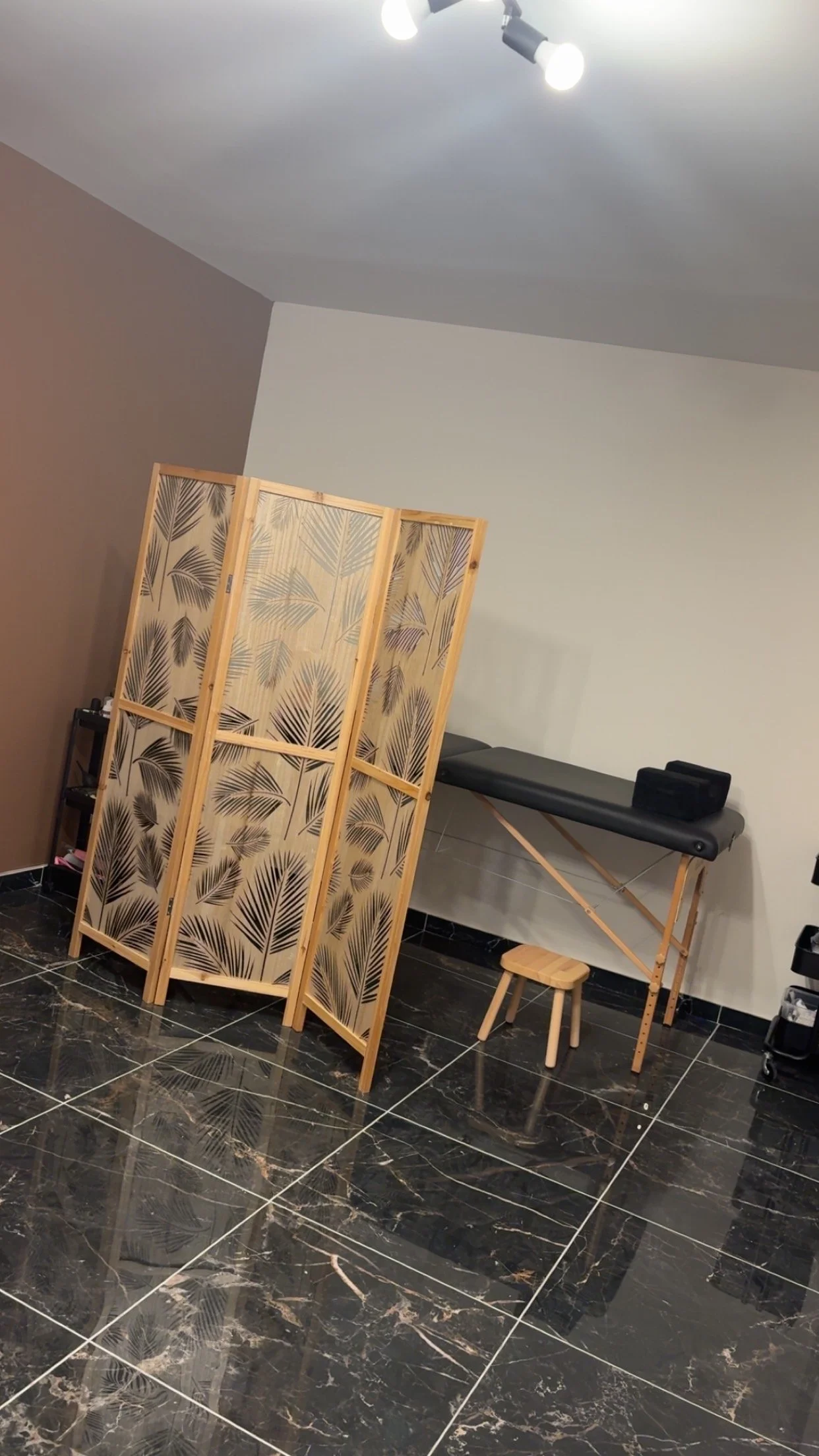 Salle de détente ou massage avec un paravent en bois avec motif de feuilles, un tableau de massage, un tabouret en bois, et un éclairage au plafond.