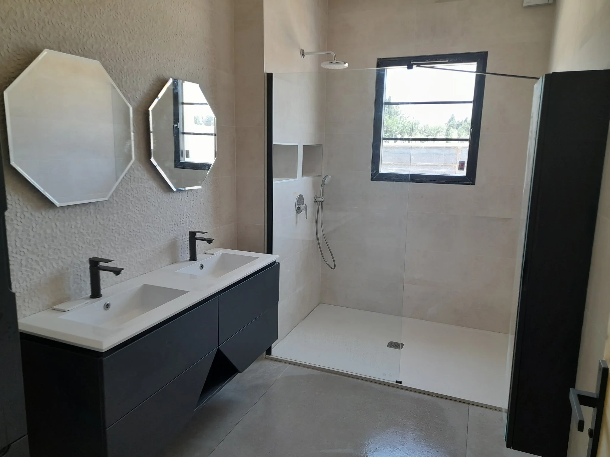 Une salle de bain moderne avec double lavabo, deux miroirs en forme hexagonale, une cabine de douche avec porte en verre, deux fenêtres pour la lumière naturelle, un sol en carrelage gris et un mur beige.