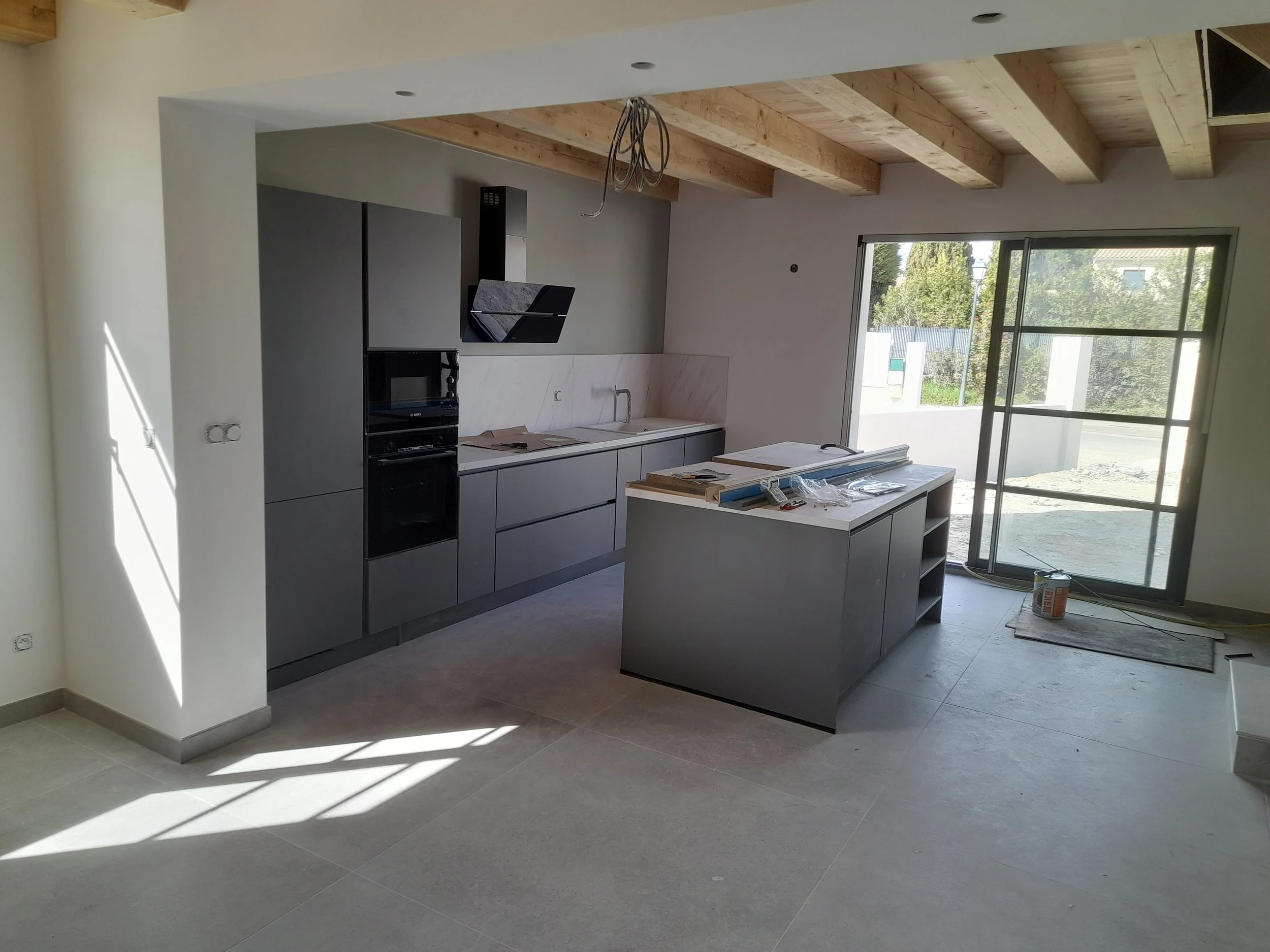 Intérieur d'une cuisine en cours de construction ou de rénovation, avec des armoires modernes de couleur grise, un plan de travail, et une grande fenêtre laissant entrer la lumière naturelle.