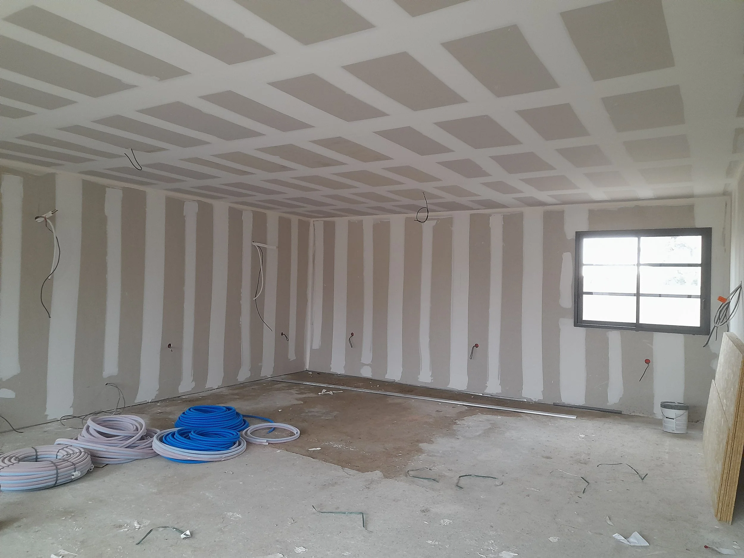 Une pièce en cours de construction avec des murs partiellement finis, des fils électriques suspendus au plafond, des bobines de câble au sol et une fenêtre à l'arrière.