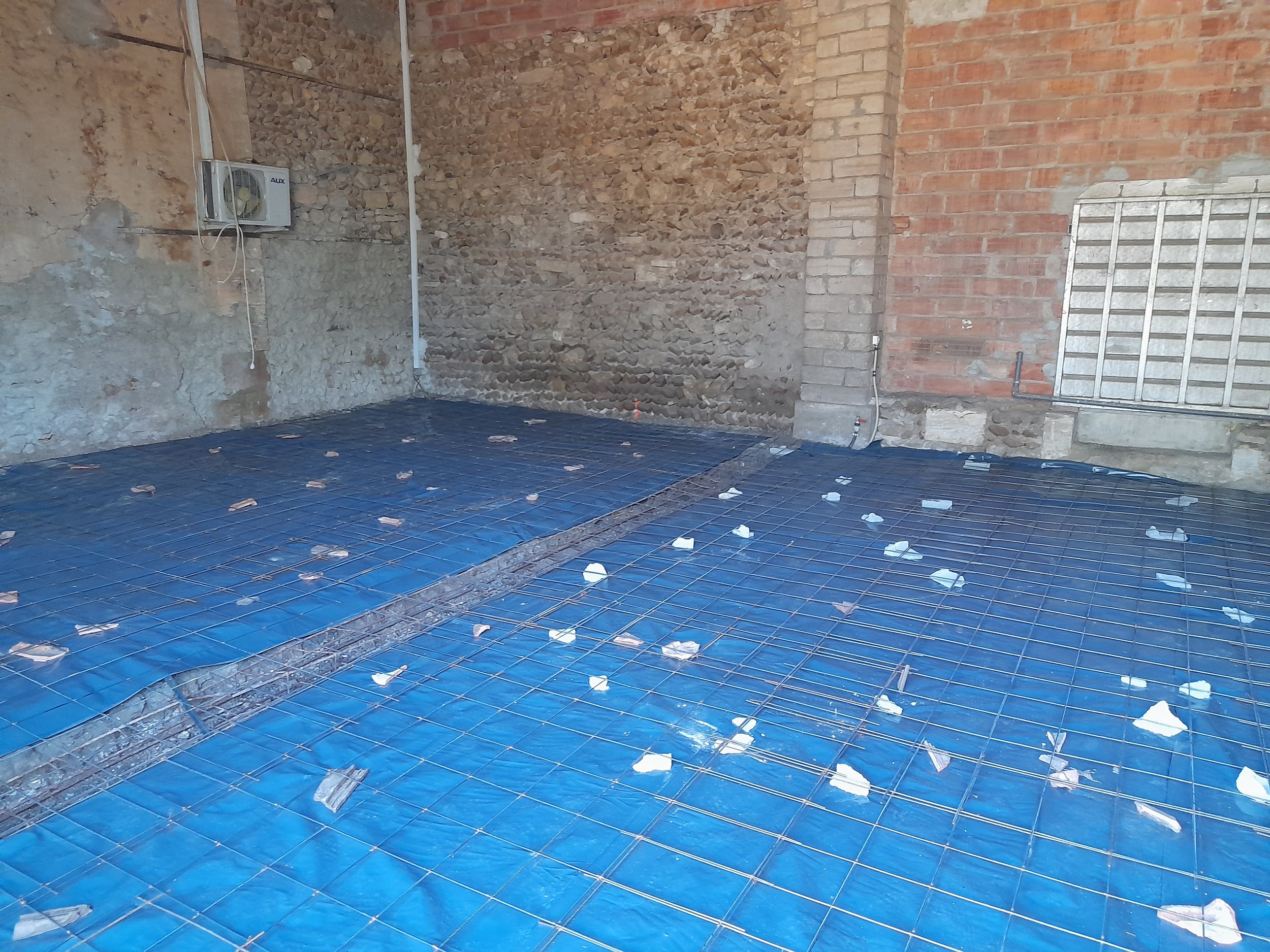 Une pièce en cours de rénovation avec un sol en grille métallique. Les murs en briques apparentes et en pierre brute, avec un panneau de grille en métal sur le mur droit. Un petit radiateur électrique est fixé à gauche, avec des fils électriques visi