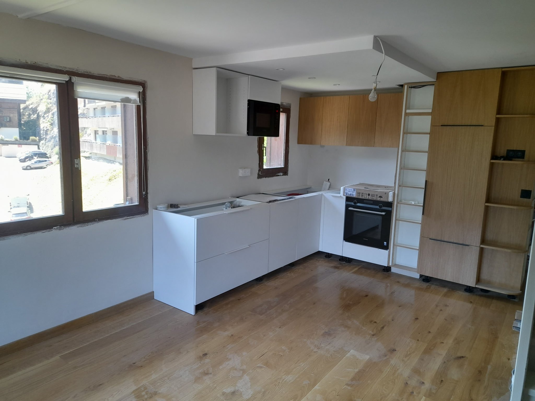 Intérieur d'une pièce en cours de rénovation avec fenêtres laissant entrer la lumière naturelle, cuisine partiellement installée avec meubles en bois clair et comptoir blanc, sol en bois.