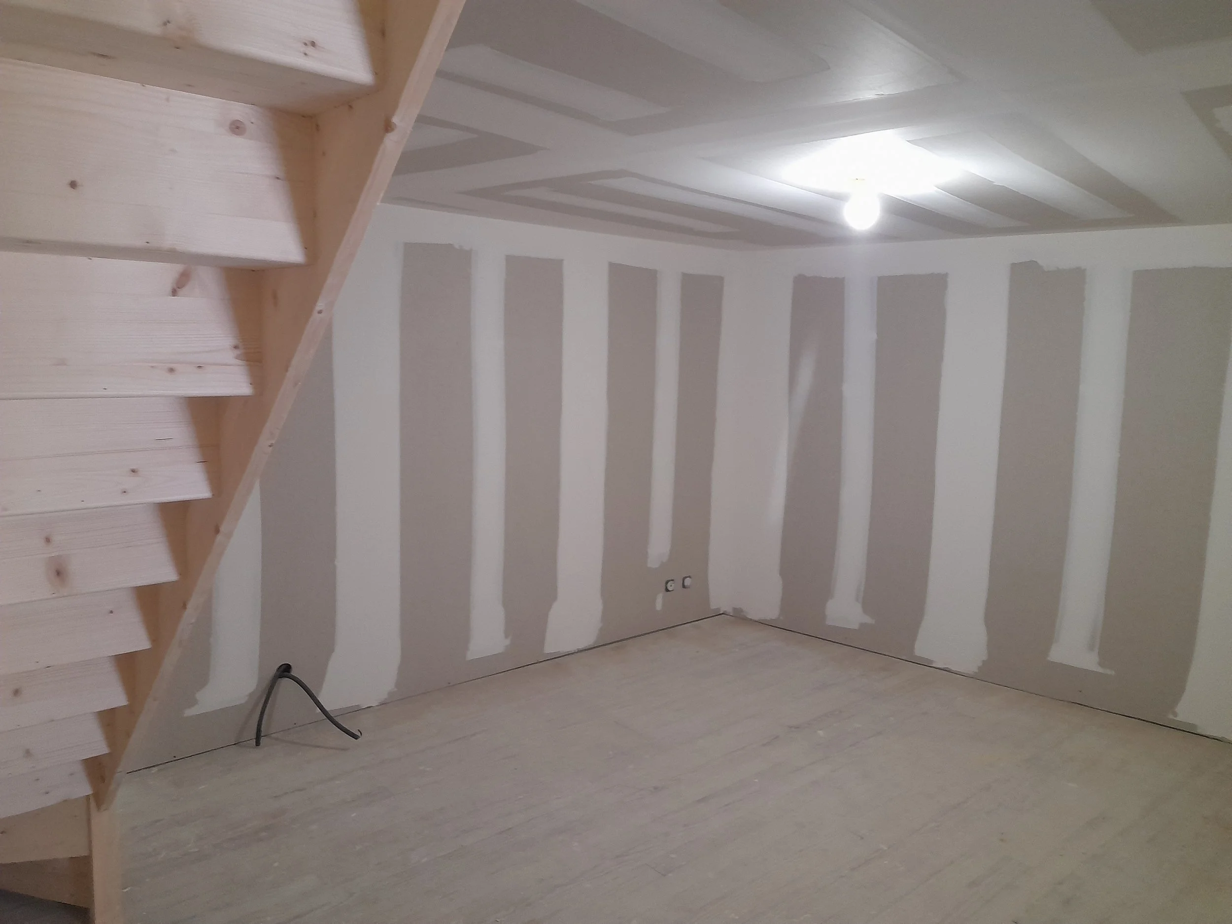 Intérieur d'une pièce en cours de rénovation avec des murs en plâtre et un plafond avec des bandes en peinture blanche. Un escalier en bois non fini est visible à gauche, et un câble électrique noir est par terre.