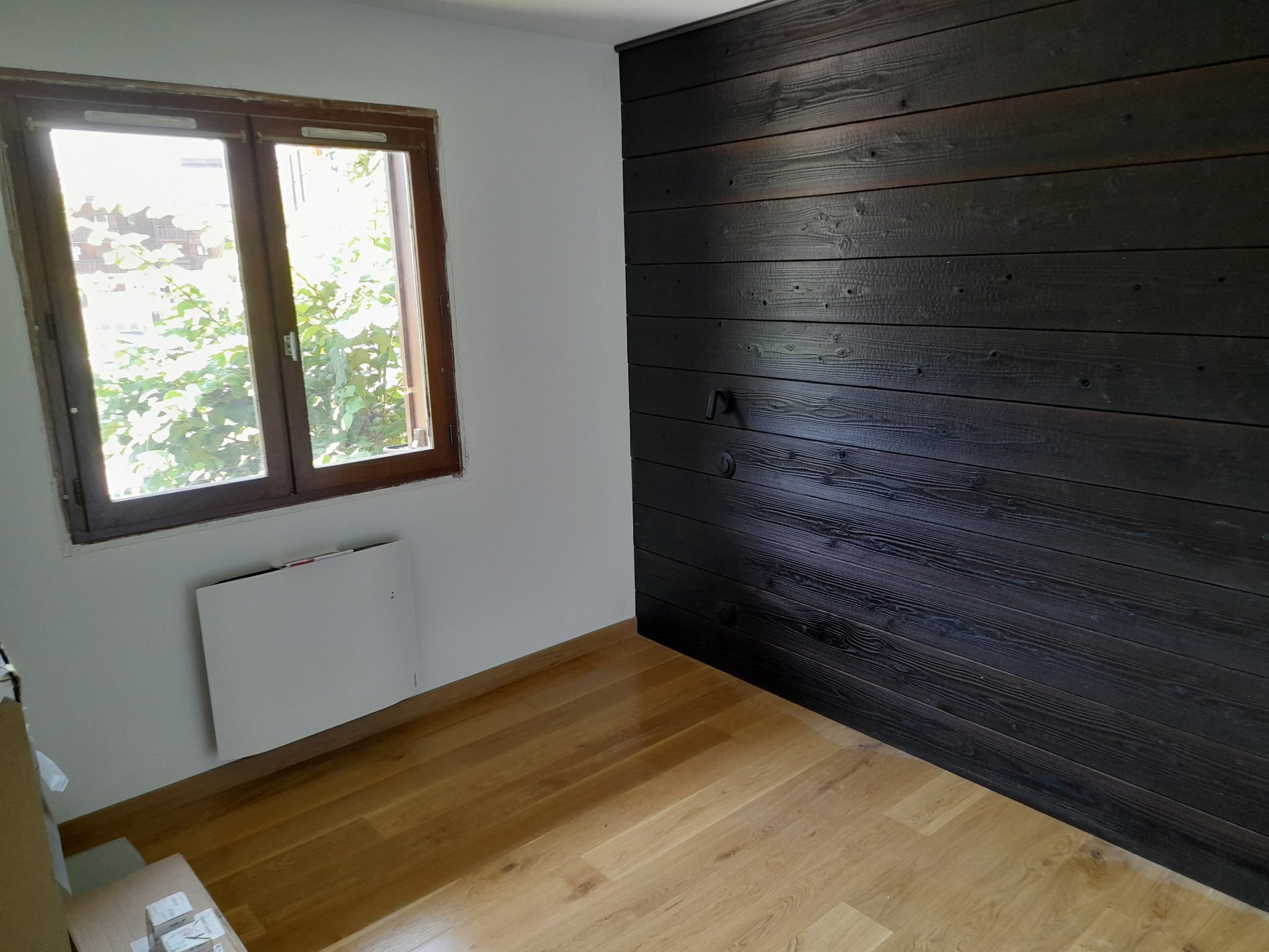 Une pièce avec une fenêtre en bois, un mur blanc, et un mur en bois foncé. Le sol est en bois clair.
