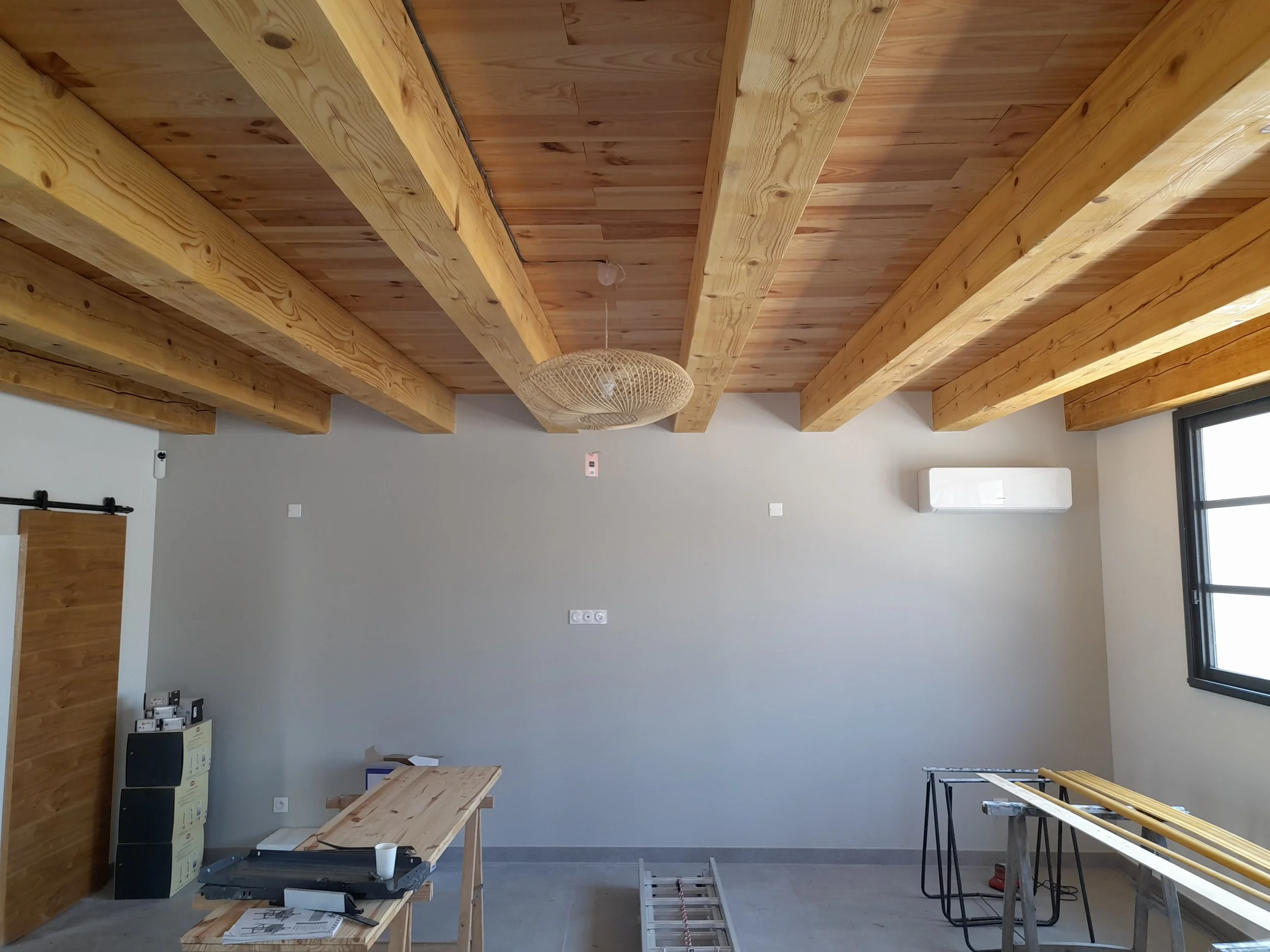 Intérieur d'une pièce en construction avec des poutres en bois apparentes au plafond, un mur gris avec une fenêtre, une table en bois, un mélange d'outils et matériaux de construction, et un climatiseur mural.