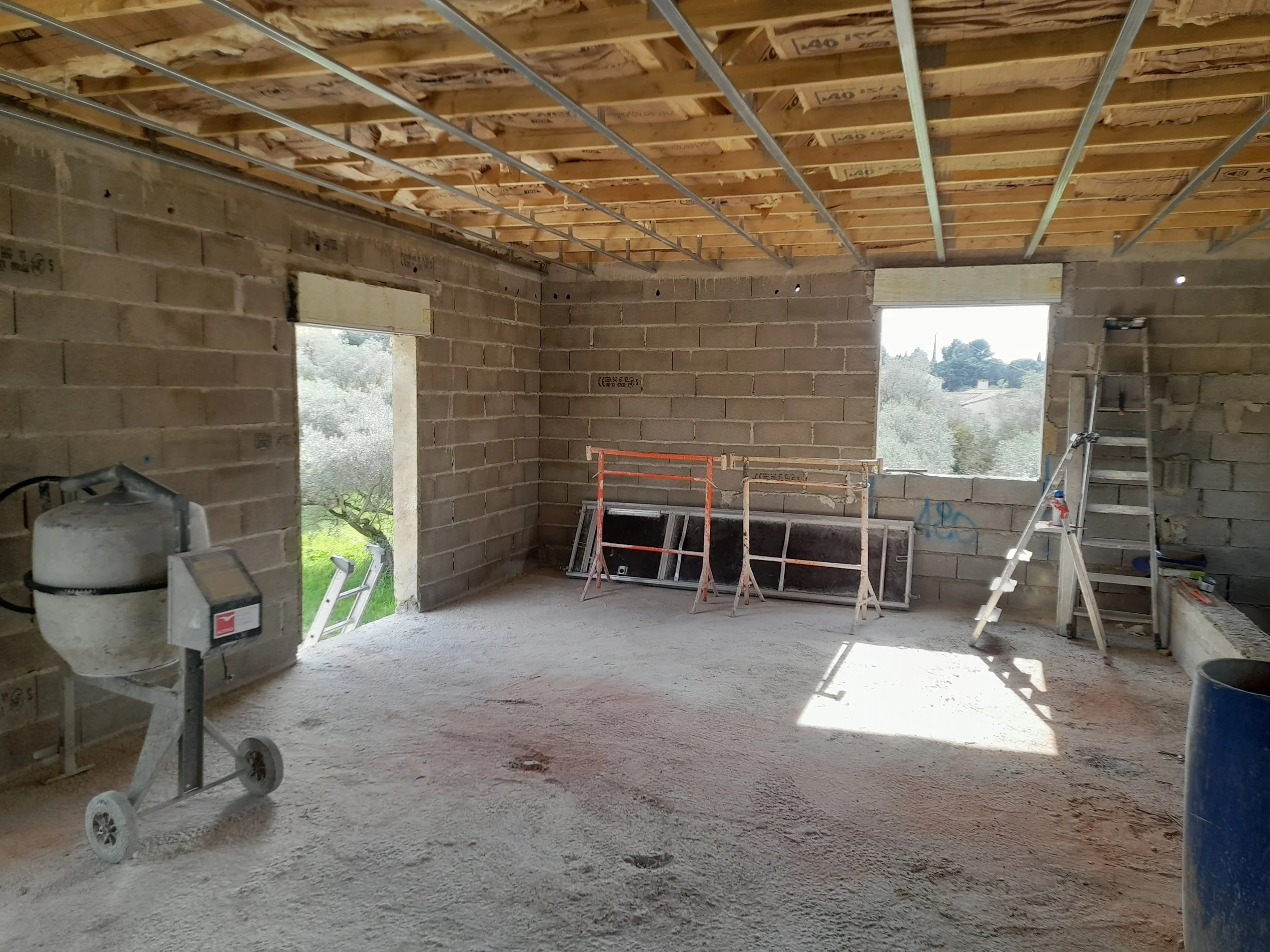 Intérieur d'une maison en construction avec murs en briques, plafond avec charpente en bois et fer, fenêtres inachevées, outils et équipement de chantier.