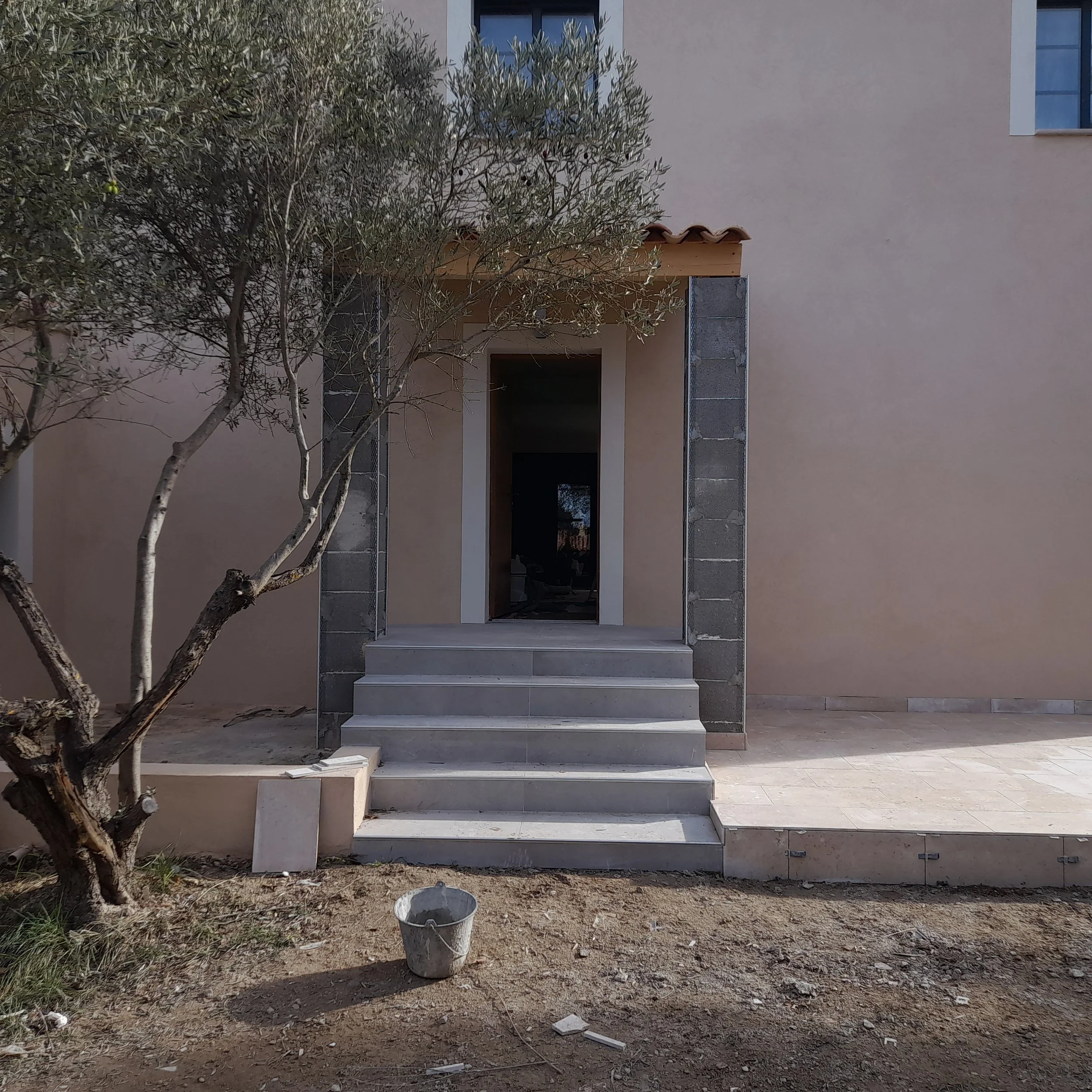 Construction d'un escalier en pierre devant une maison neuve avec un arbre à gauche.