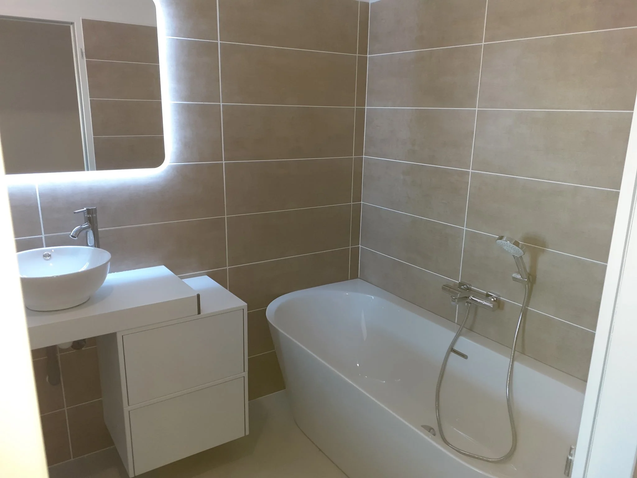 Intérieur d'une salle de bain moderne avec une baignoire, un lavabo, un miroir lumineux, et des murs carrelés en beige et gris.
