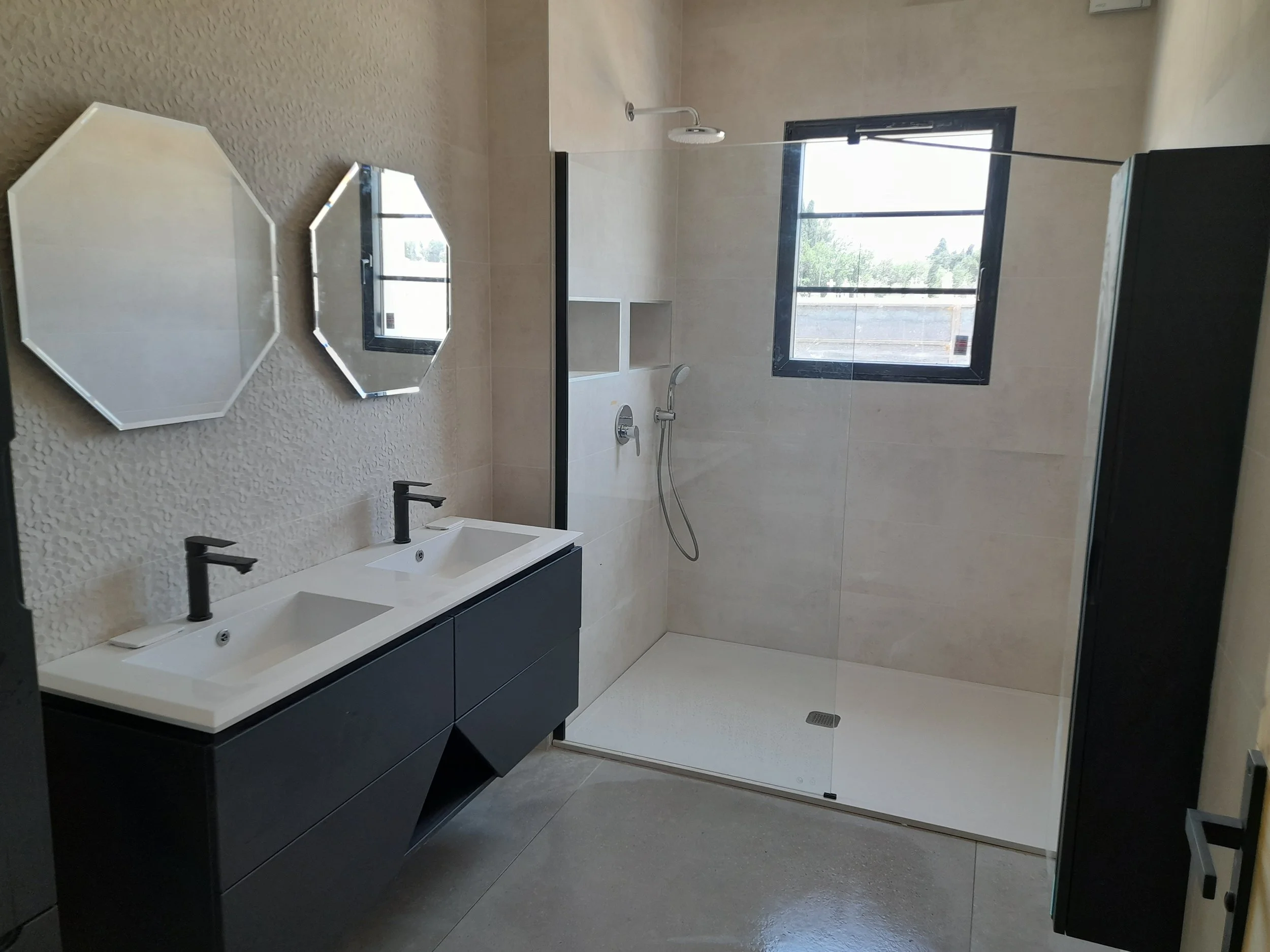 Salle de bain moderne avec double vasque, miroirs octogonaux, douche avec porte en verre, fenêtre, et meubles noirs.