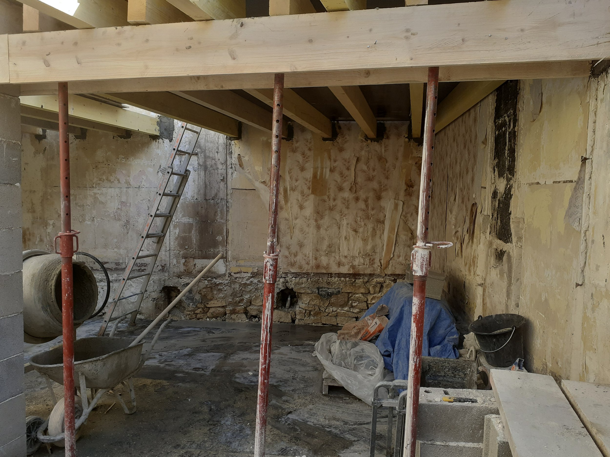 Construction intérieure en cours avec échafaudages en métal, escabeau, tuyaux, sacs de matériaux, murs en rénovation et sol en terre ou béton.