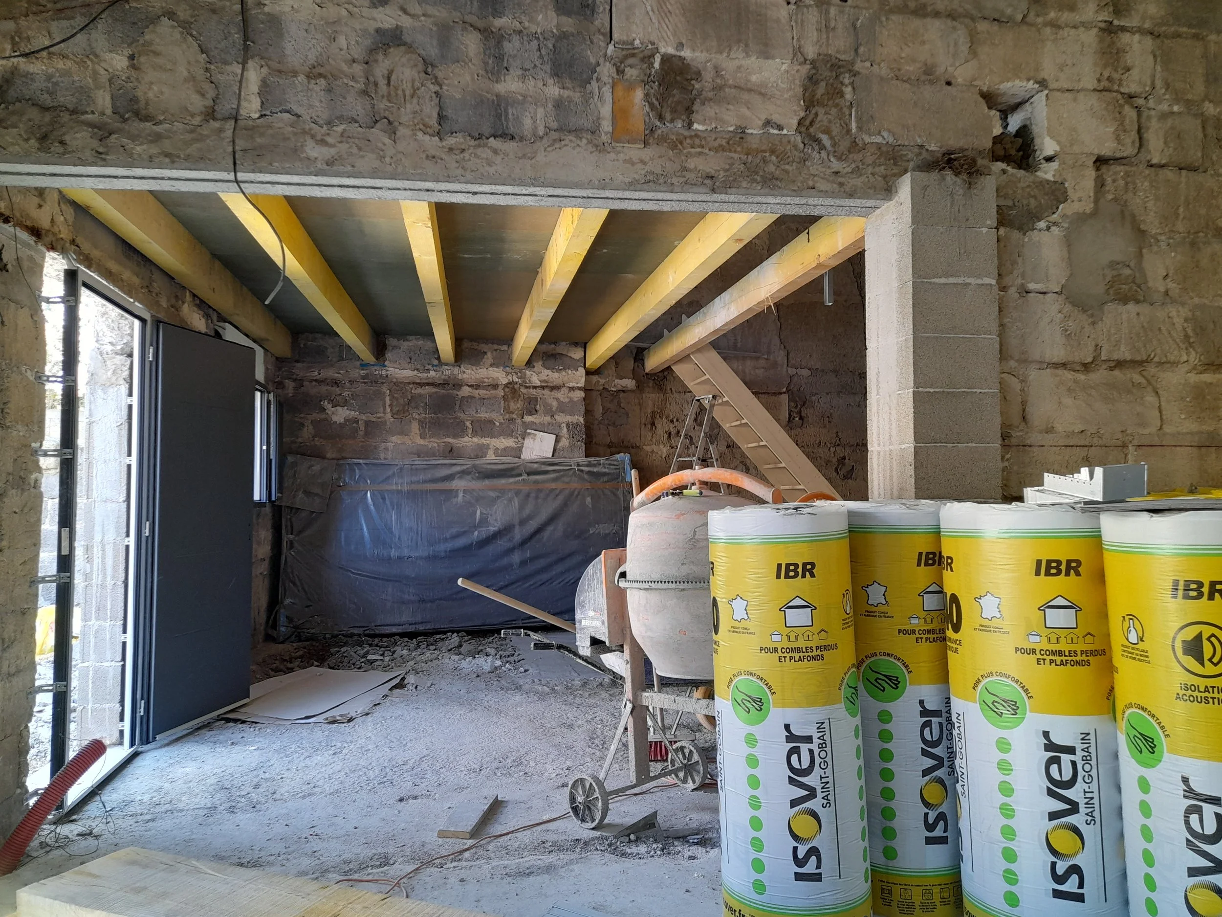 Construction intérieure en cours avec murs en pierre et sol en béton, installation de plafonds en bois, palettes de matériaux d'isolation et chauffage dans un espace en rénovation.