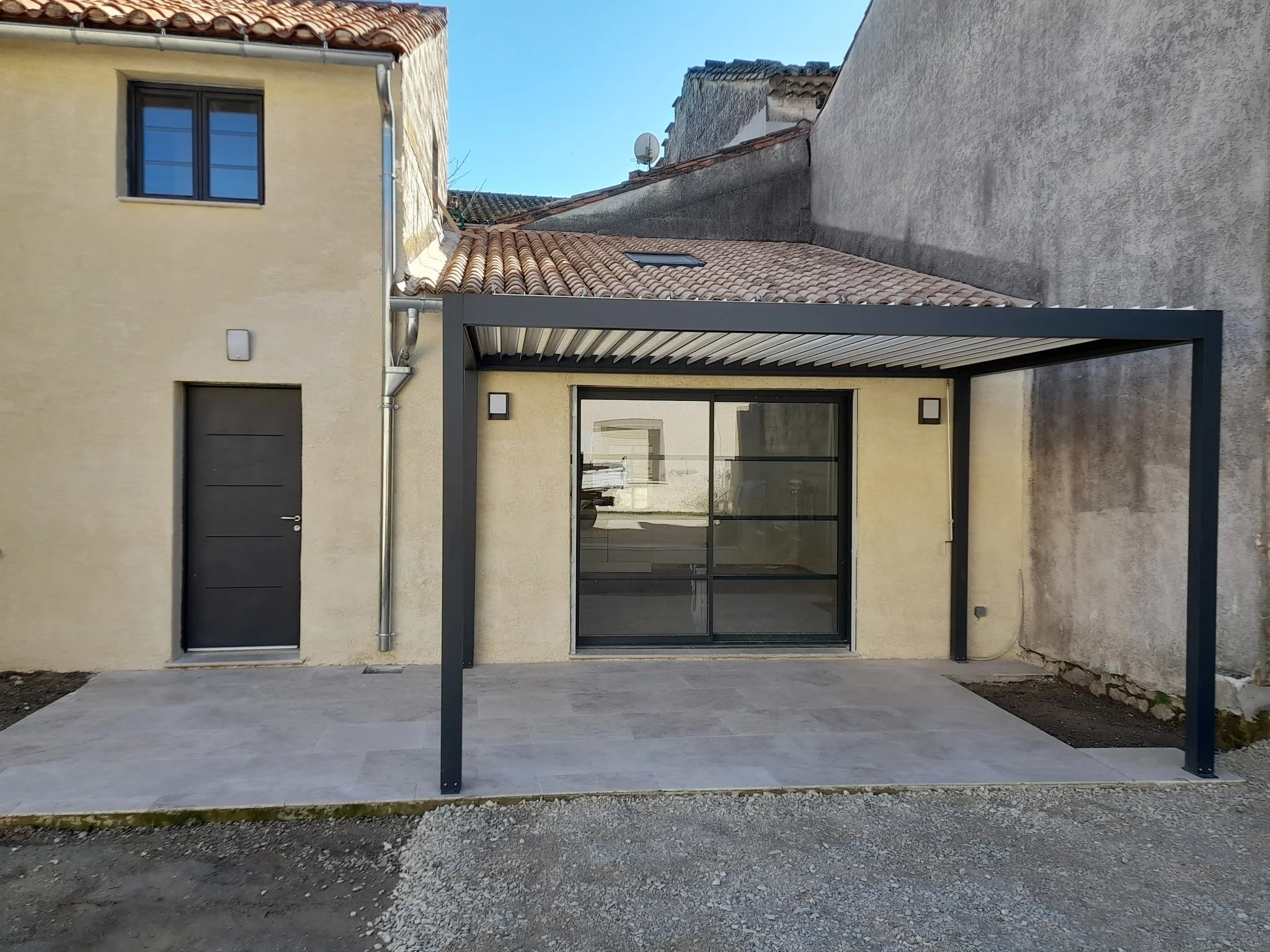 Une résidence moderne avec une façade jaune, une grande porte noire, un porte-fenêtre en verre, et une structure en métal sombre à l'extérieur, sous un ciel clair.