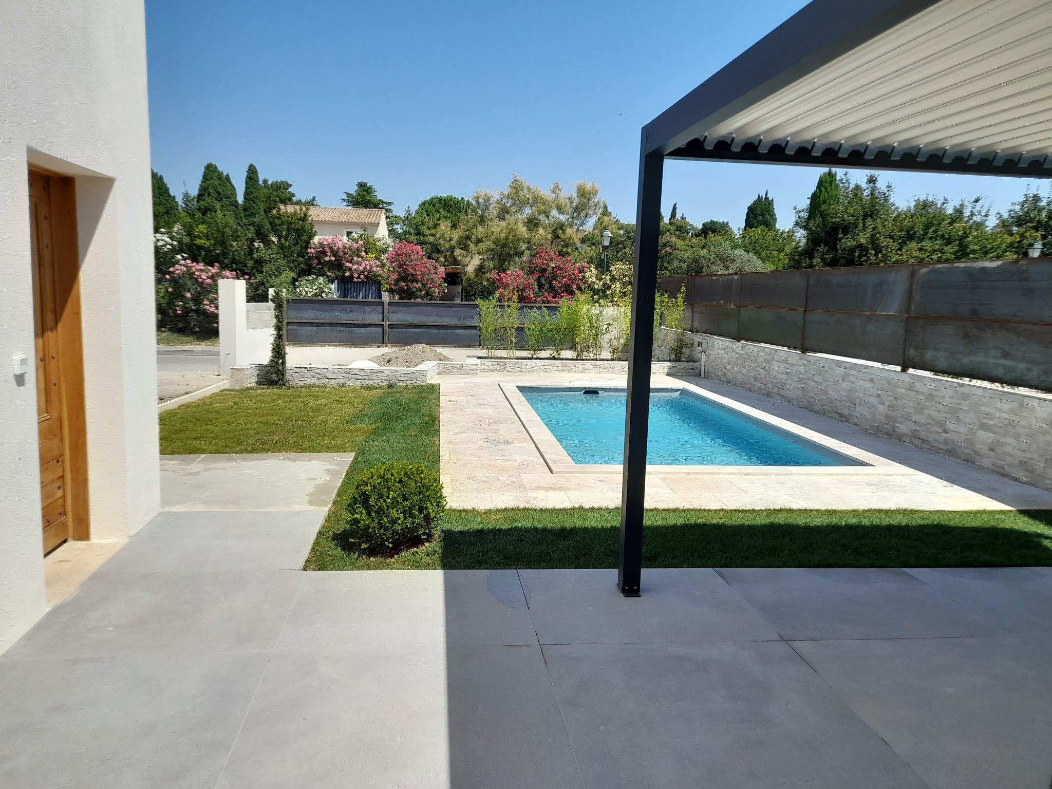 Vue d'une cour arrière avec piscine, mur de clôture, jardin, arbres et un ciel clair.