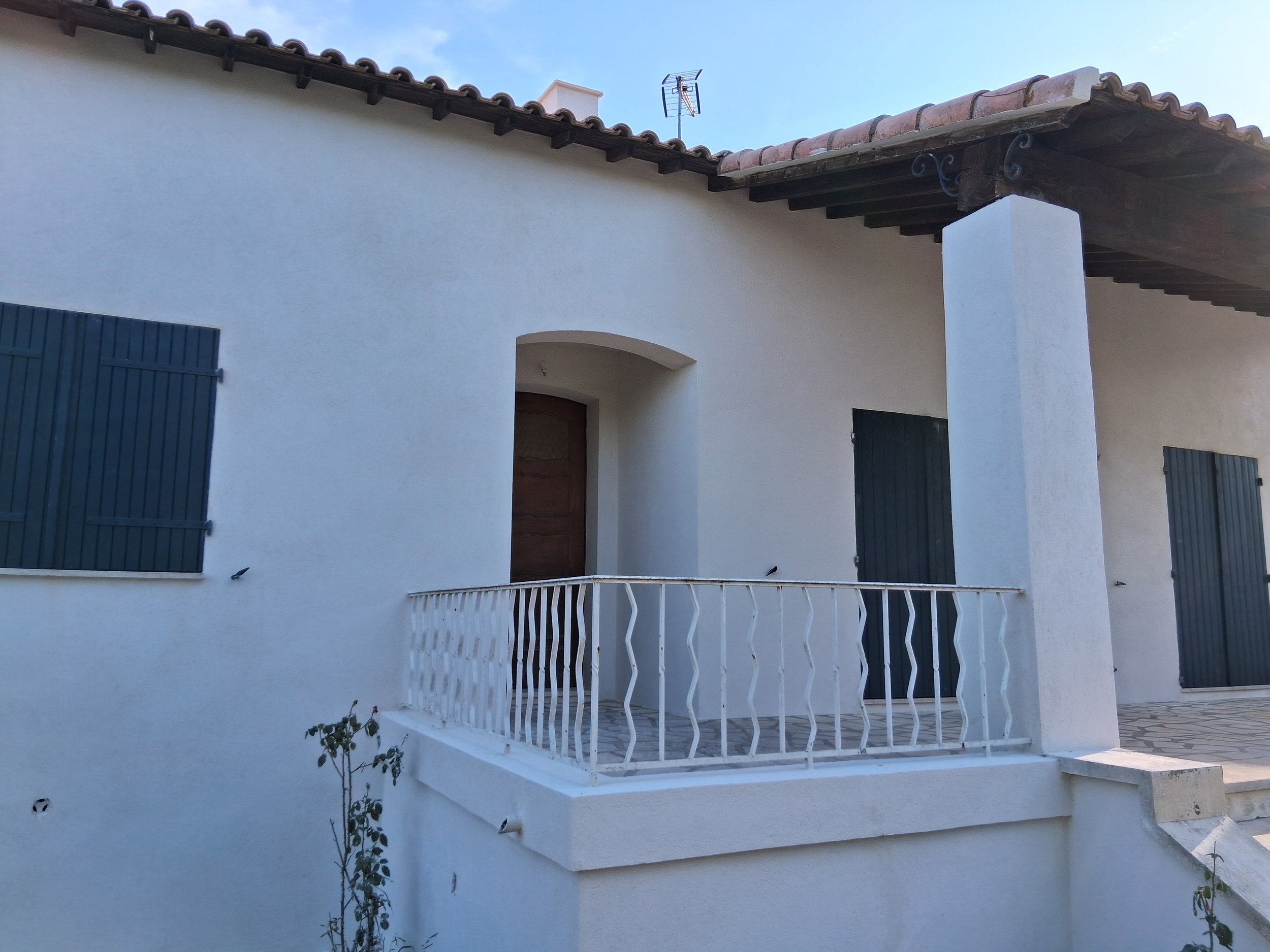 Façade d'une maison blanche avec un balcon en fer blanc, volets bleus et un toit en tuiles rouges. Un ciel bleu en arrière-plan.