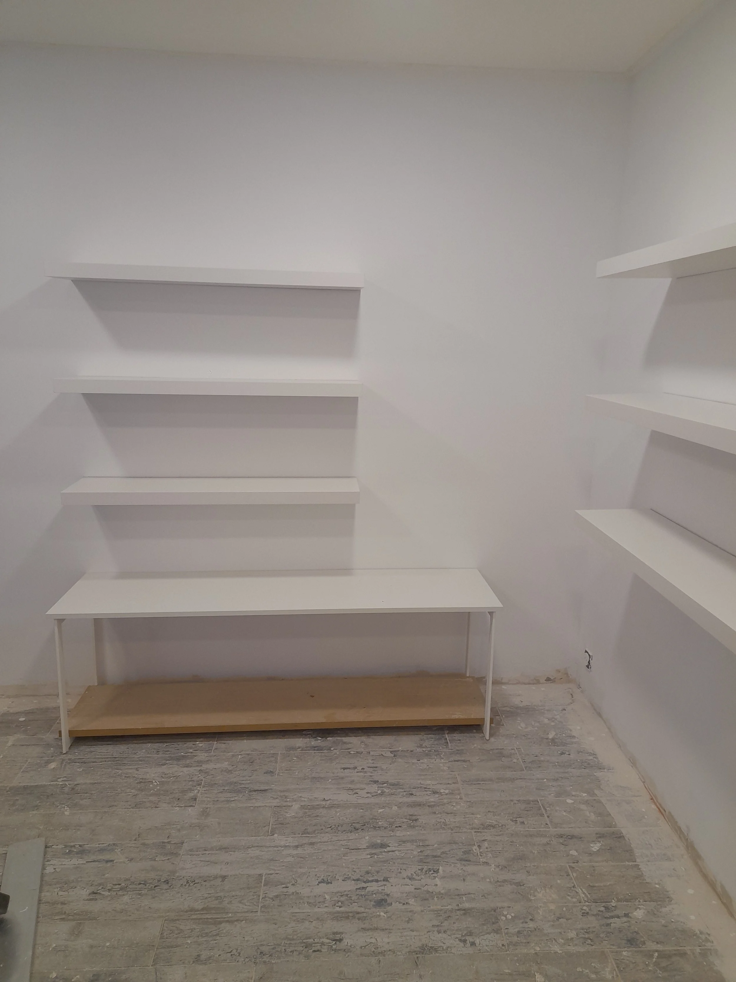 Une pièce en cours de rénovation avec des étagères blanches fixées à un mur blanc, une table en bois avec un cadre métallique en dessous, le sol en carrelage gris, et un mur avec une prise électrique.