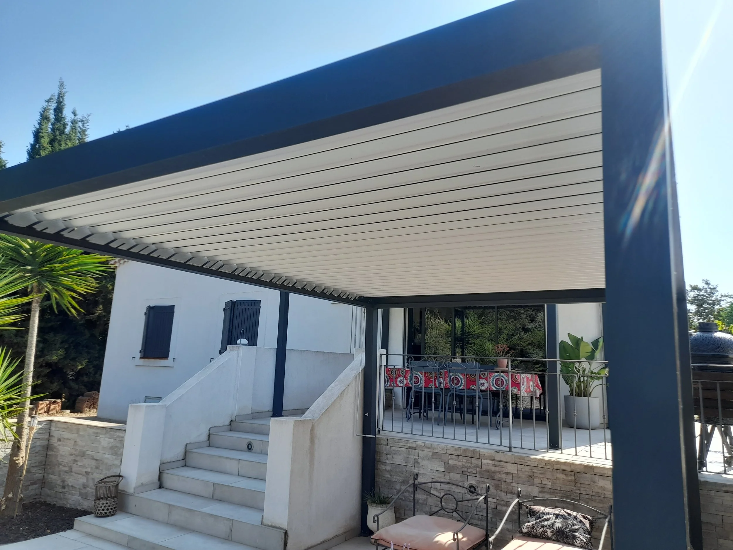 Maison moderne avec une terrasse couverte, escaliers en pierre et mobilier extérieur, entourée de végétation sous un ciel dégagé.