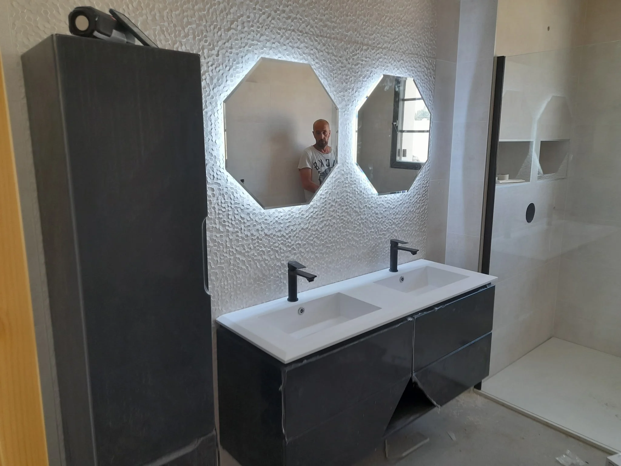 Un miroir hexagonal dans une salle de bain, reflet d'un homme avec une moustache et un t-shirt blanc, placé au-dessus de deux lavabos noirs avec robinet centrée.