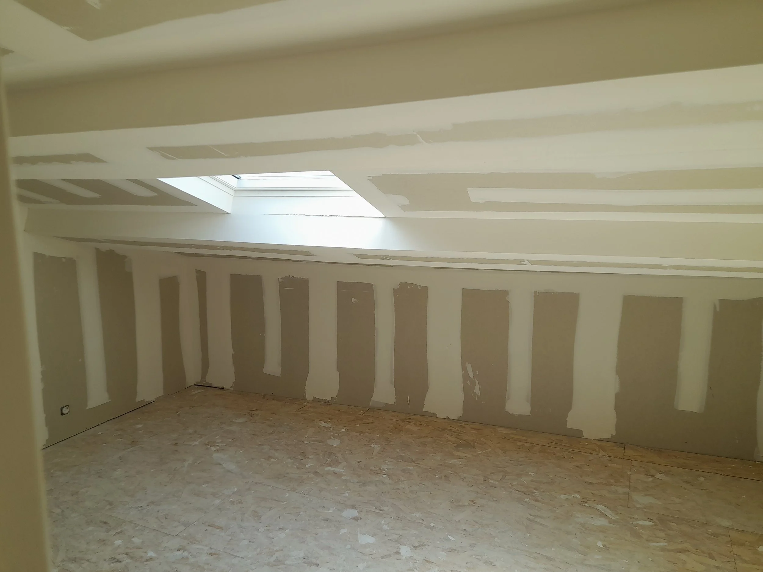 Une pièce à l'état de construction avec des murs en placo en cours d'installation, un plafond incliné avec une fenêtre de toit laissant entrer la lumière, le sol en panneau de bois. 