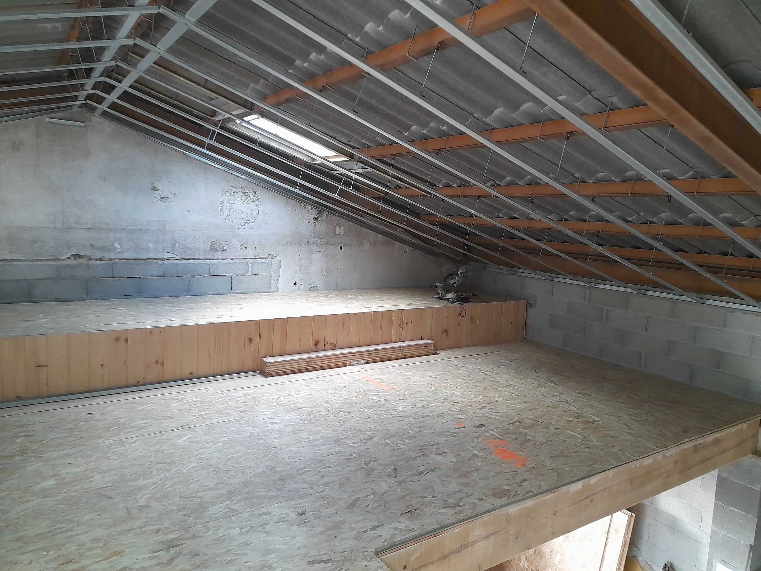 Intérieur d'un bâtiment en construction avec plafond en béton, poutres en bois, murs en brique ou en béton, et sol en planches de bois.