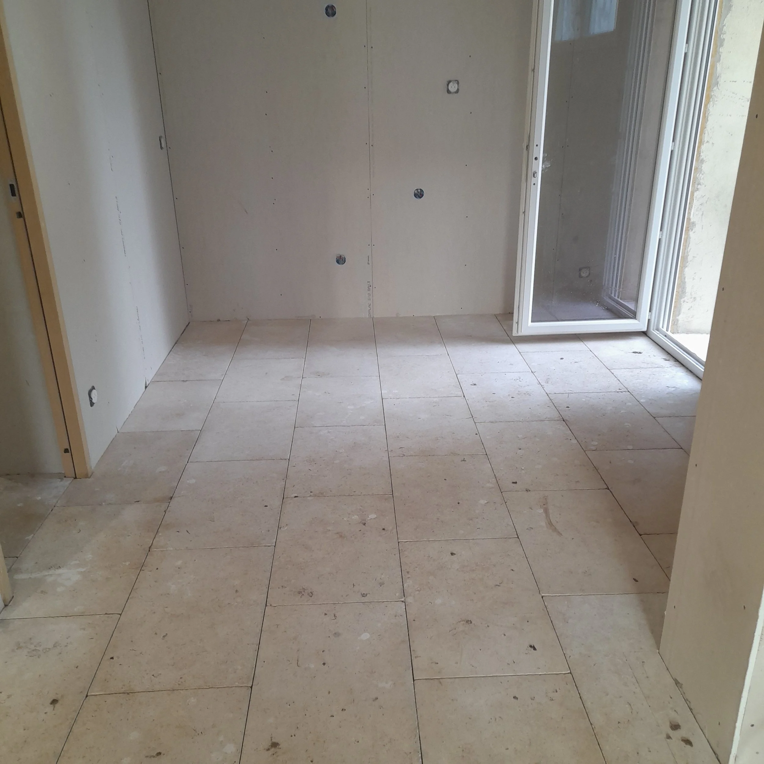 Pièce en cours de rénovation avec murs en plaques de plâtre, prises électriques et sol carrelé, vue depuis une porte.